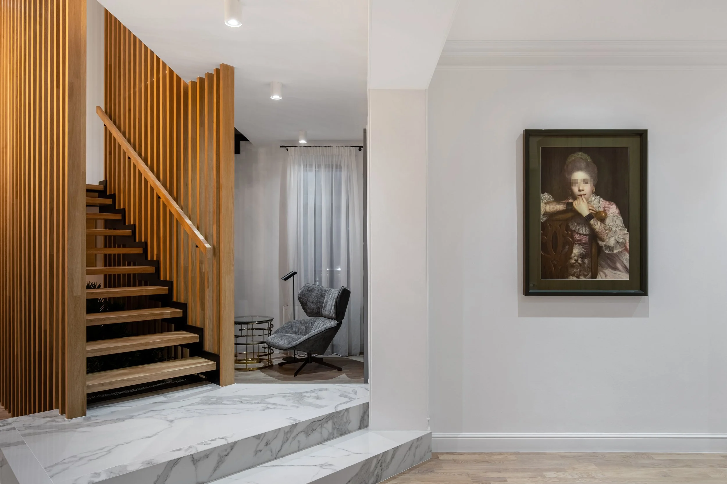 Modern interior with a staircase featuring wooden slats, a gray lounge chair, a small side table, a floor lamp, and a framed painting of a young girl on a white wall.