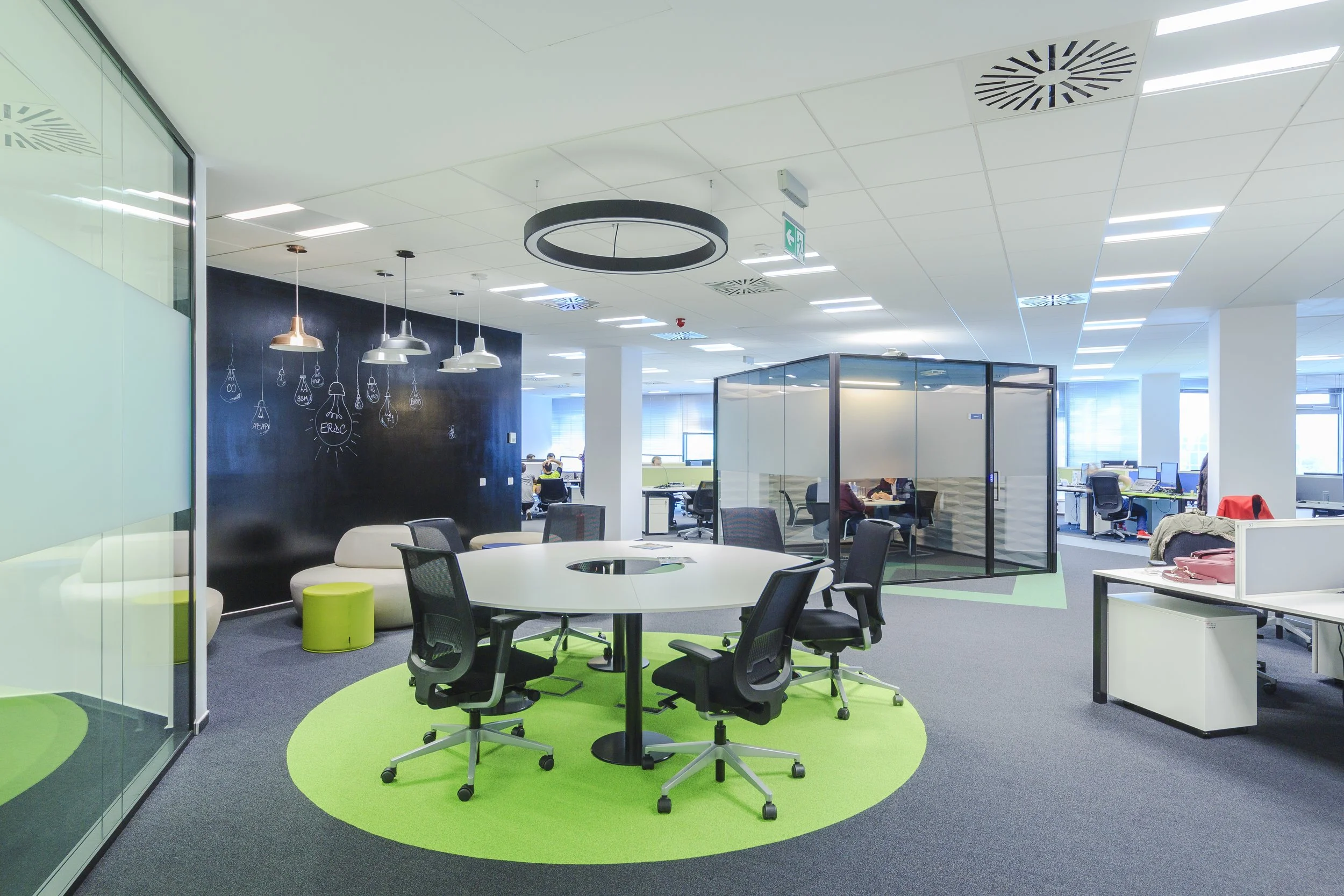 Modern open-concept office space with a round table surrounded by black office chairs on a green circular rug, a small white sofa with green stools, glass-walled meeting room, and workstations in the background.