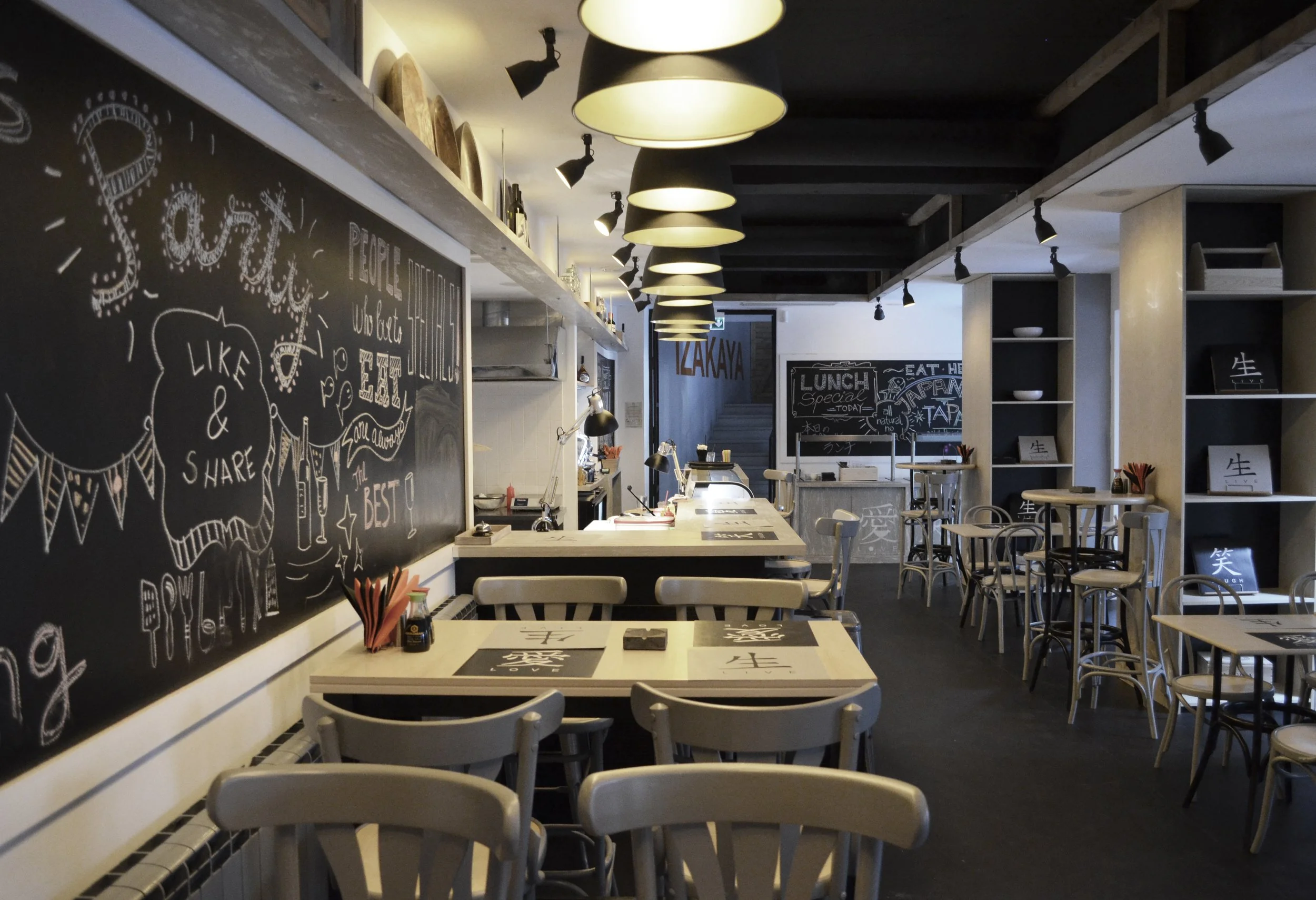 Interior of a modern restaurant with wooden tables, chairs, blackboards with chalk writings, and shelves with decorative items, lit by pendant lights.