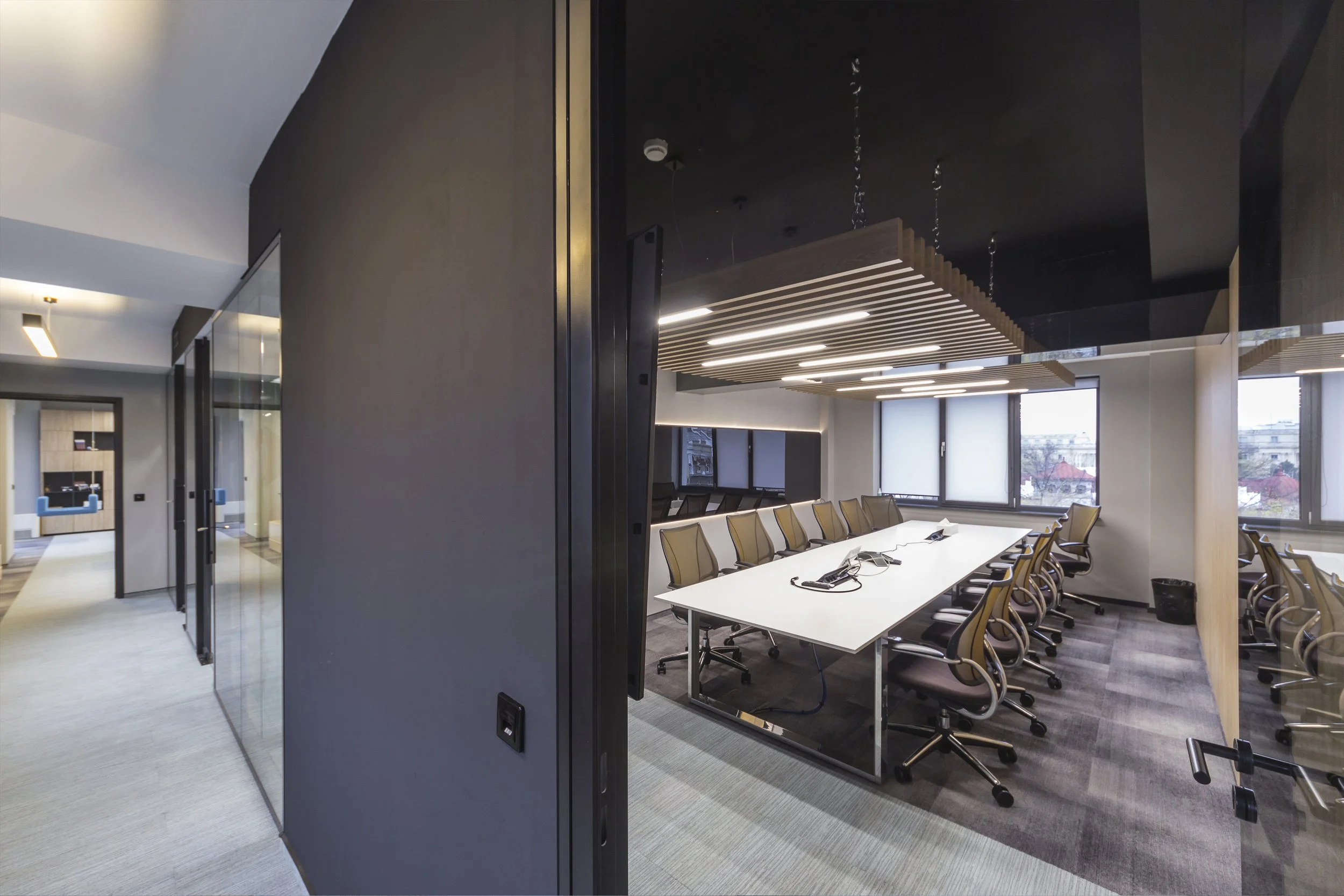 Modern conference room with a long white table surrounded by black chairs, large windows, and contemporary ceiling lighting.
