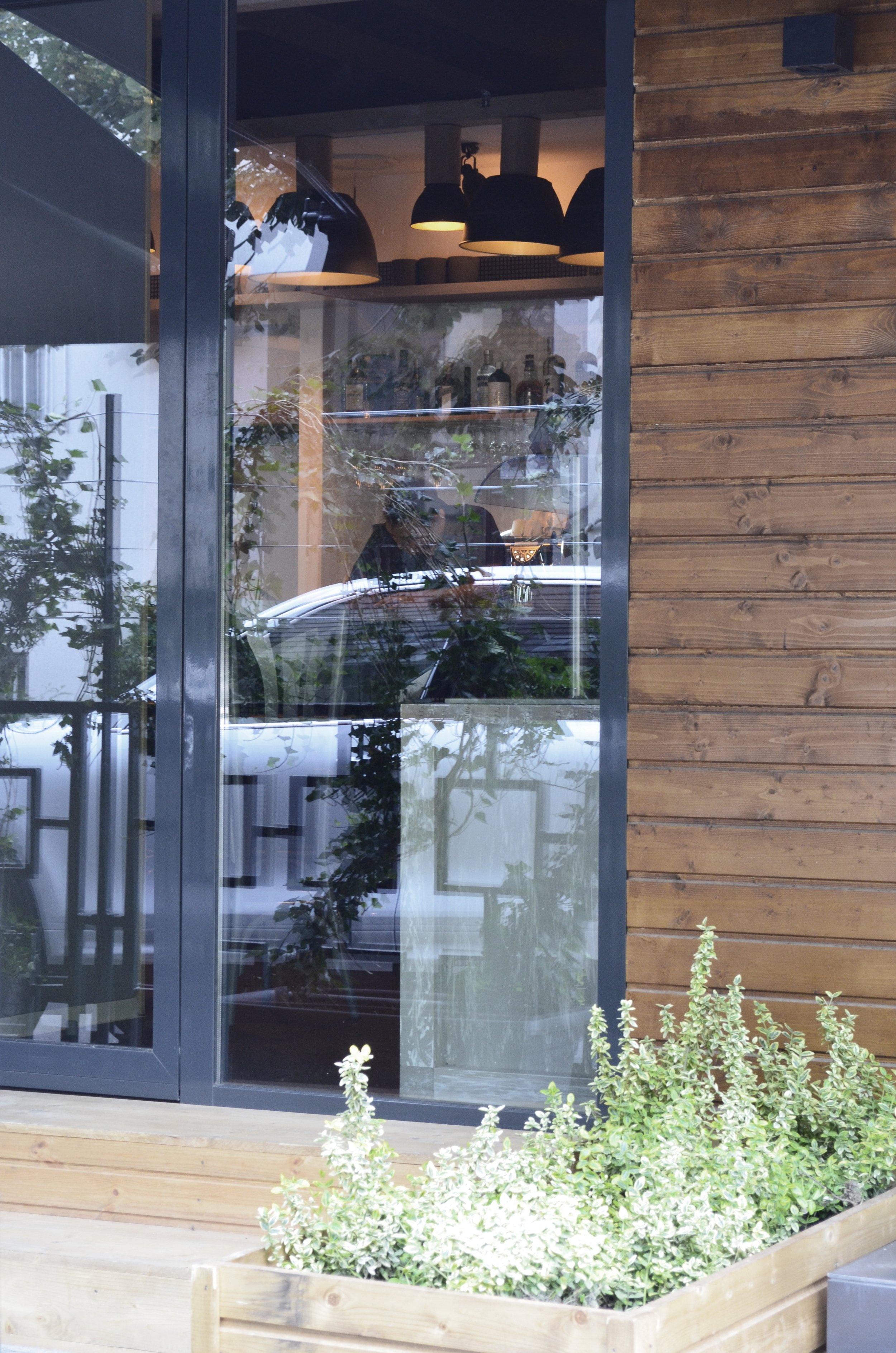 A window with a view into a cafe or restaurant, showing a person working inside, LED hanging lamps, bottles on bottles, plants outside, and a wooden exterior wall.