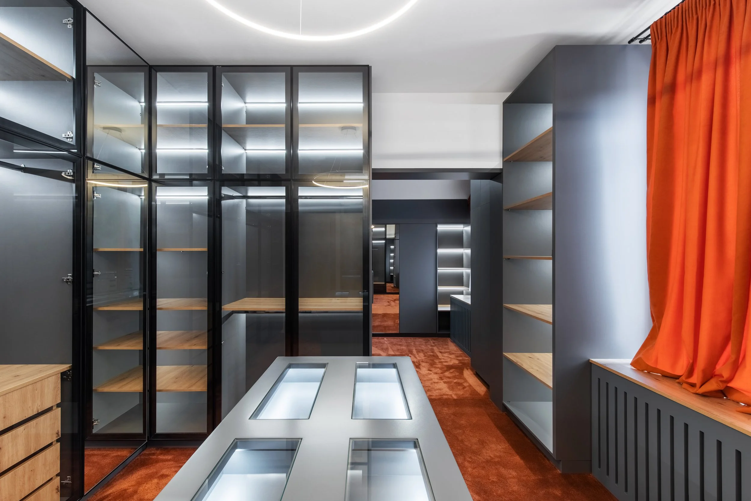 An modern room with gray walls and black framed glass cabinets with wooden shelves, an orange curtain on a window, and a white table with glass insets. The room has a warm, contemporary design with orange carpeting.