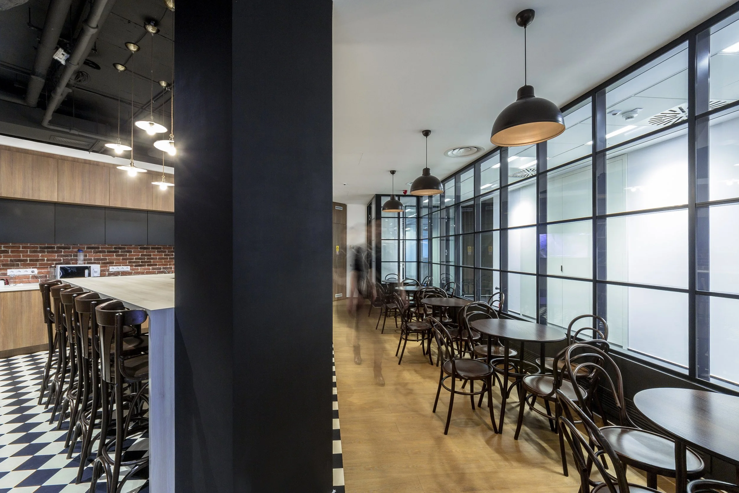 Modern cafe with black and wood accents, round tables with wooden chairs, large glass windows, black pendant lights, checkered floor near the counter, and a blurred person walking.