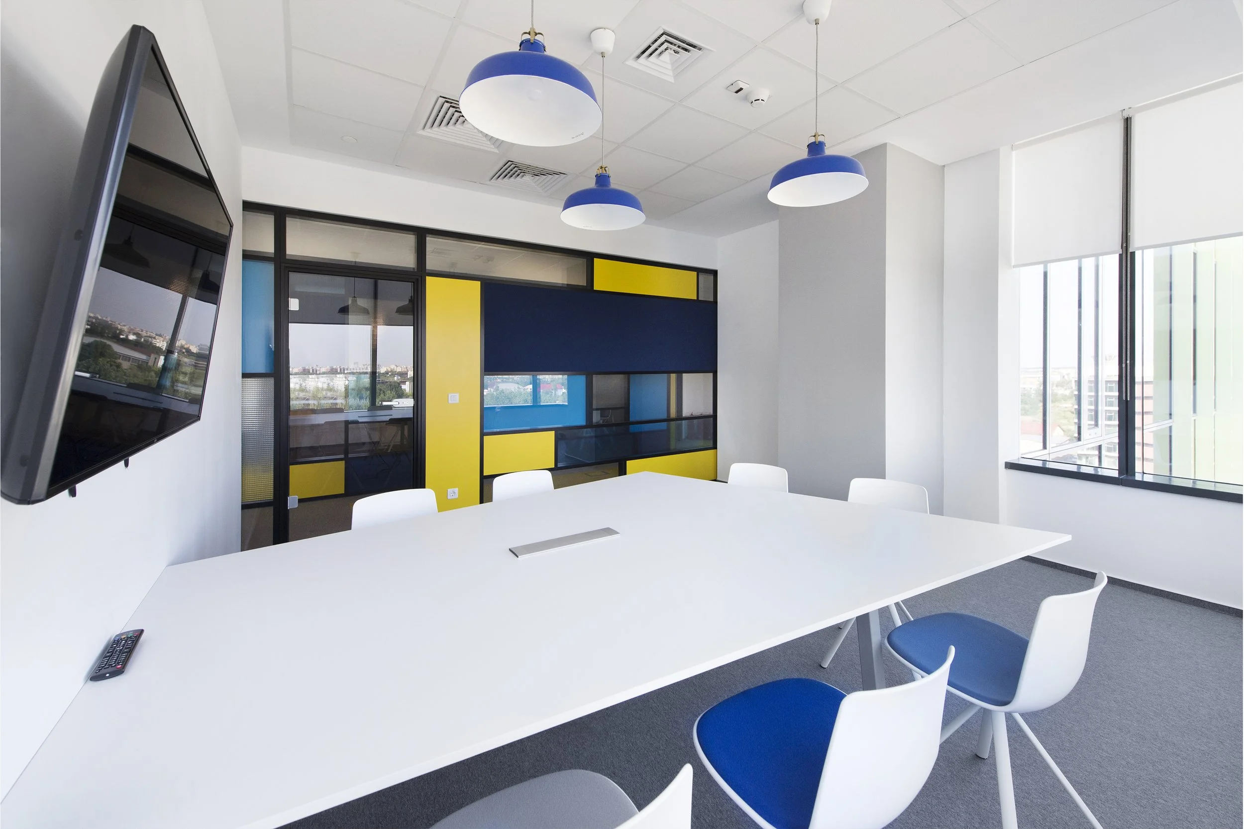 Empty modern conference room with white table, white and blue chairs, large windows, and colorful wall decor featuring blue, yellow, and black panels.