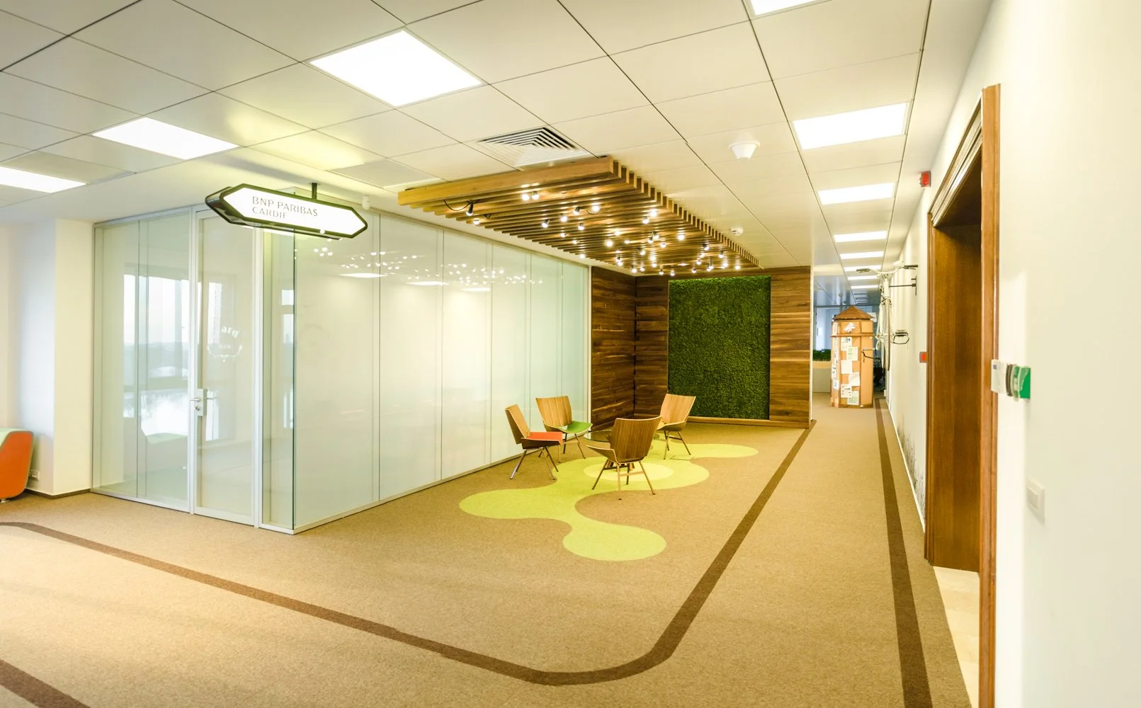 Modern office corridor with wooden wall decor, ceiling lights, and a glass-walled room labeled 'BNP Paribas Cardiff'. There is a small seating area with four chairs and playful yellow carpet patterns. Cardboard boxes are stacked toward the end of the corridor.