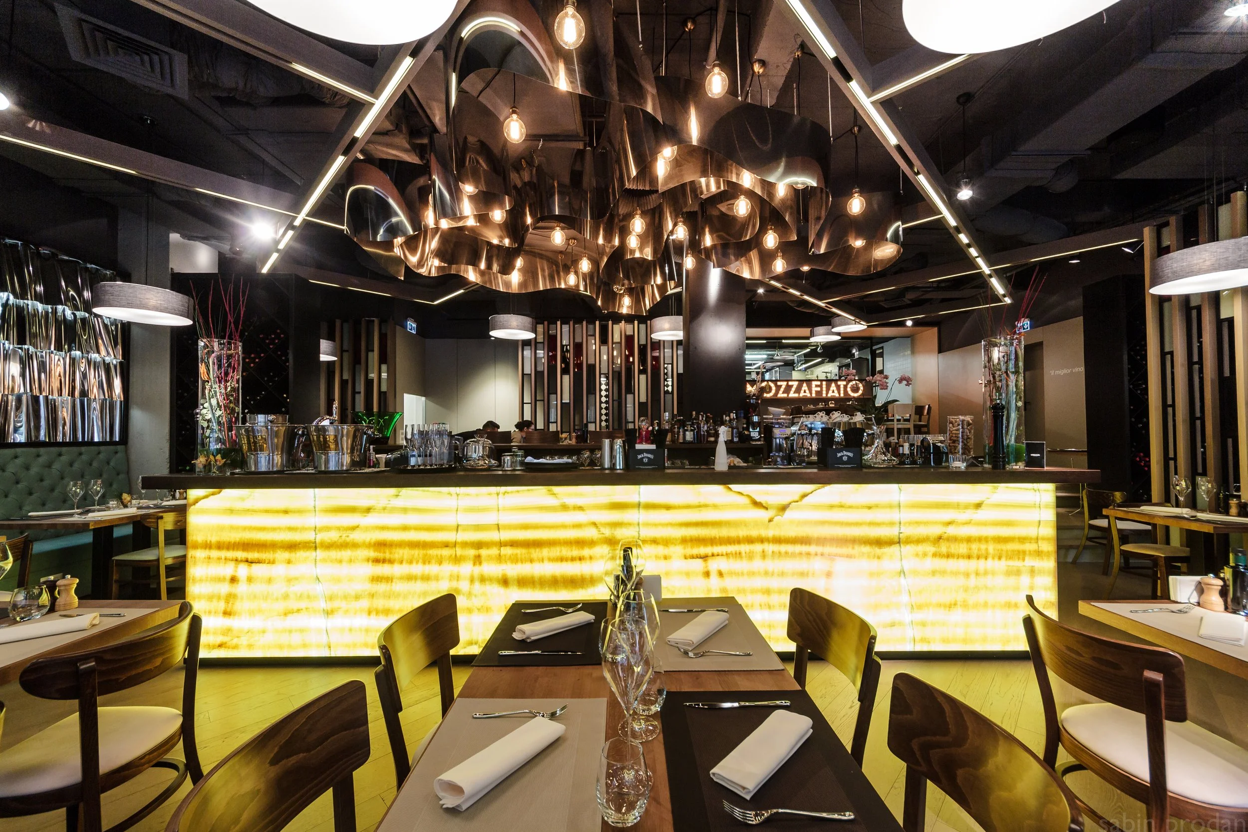 A modern restaurant interior with a brightly lit yellow bar counter, wooden chairs with white seat cushions, tables set with napkins, glasses, and cutlery, decorative lighting fixtures, and a sign that reads 'PIZZAFIATO' in the background.