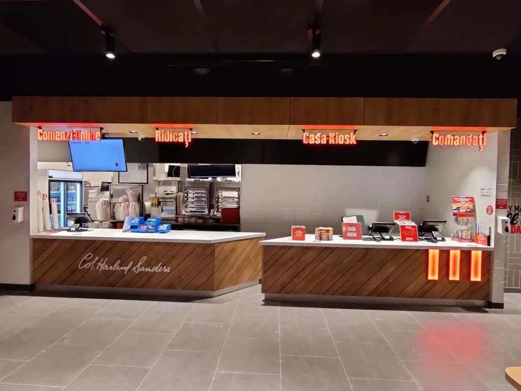 A fast food takeout counter with signs in Spanish and English, including options for online ordering, pickup, and drive-thru, featuring drink dispensers, a cash register, and menu displays.