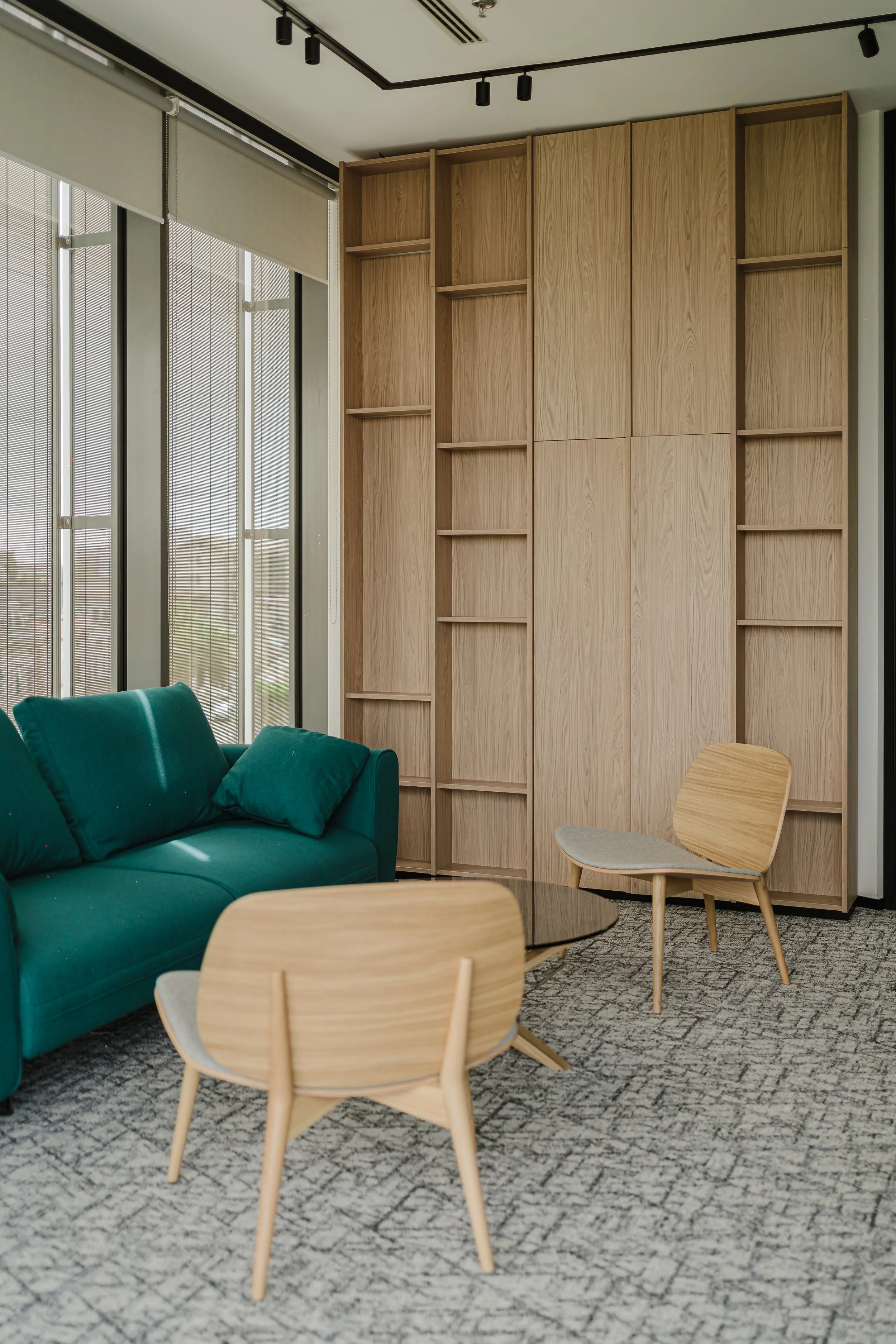 Modern office lounge area with a teal sofa, wooden chairs, a round glass coffee table, and a wooden bookshelf against a large window with blinds.