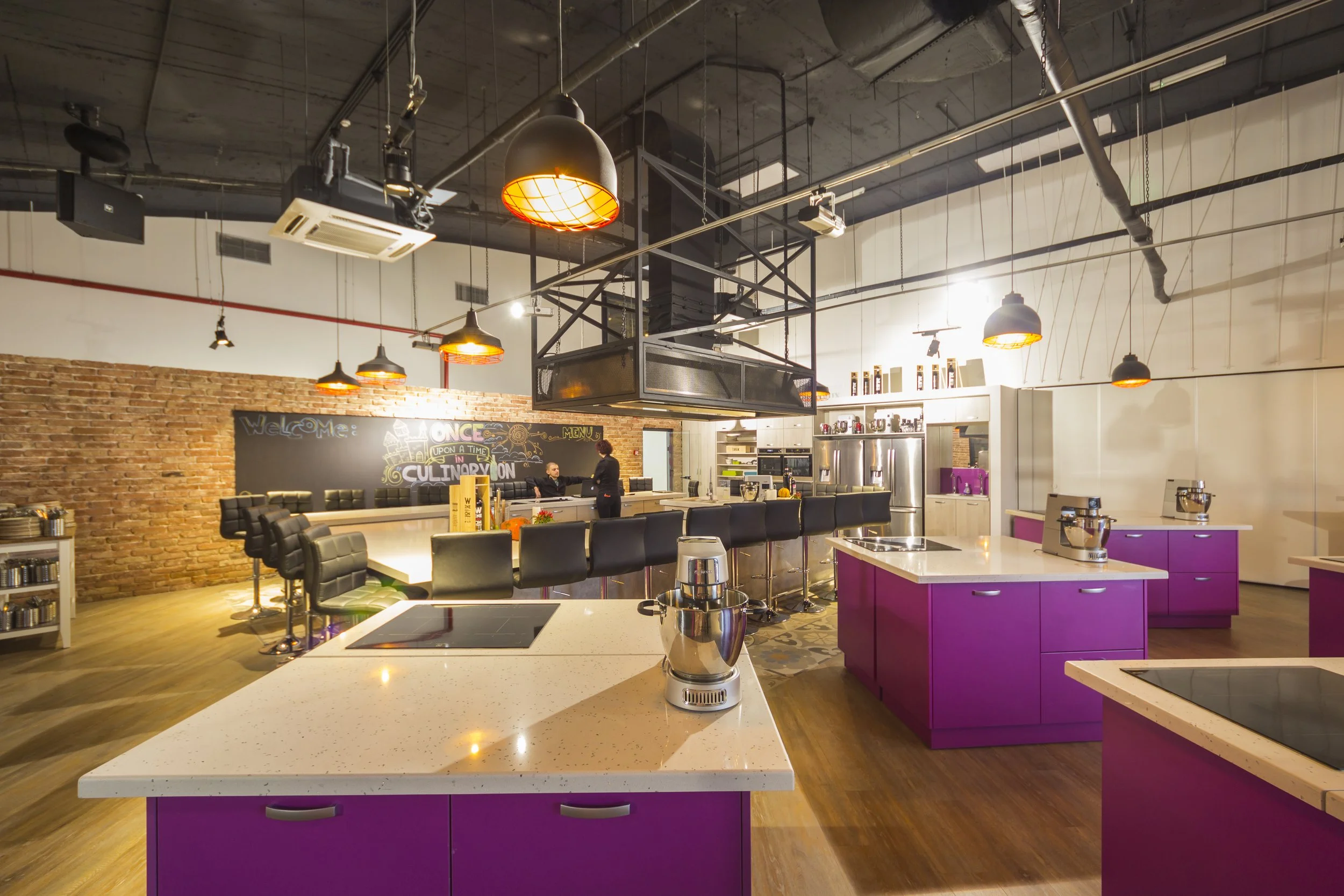 Modern industrial kitchen with purple cabinets and black chairs, large island with coffee makers, exposed brick wall, chalkboard with welcome message, and staff preparing food.