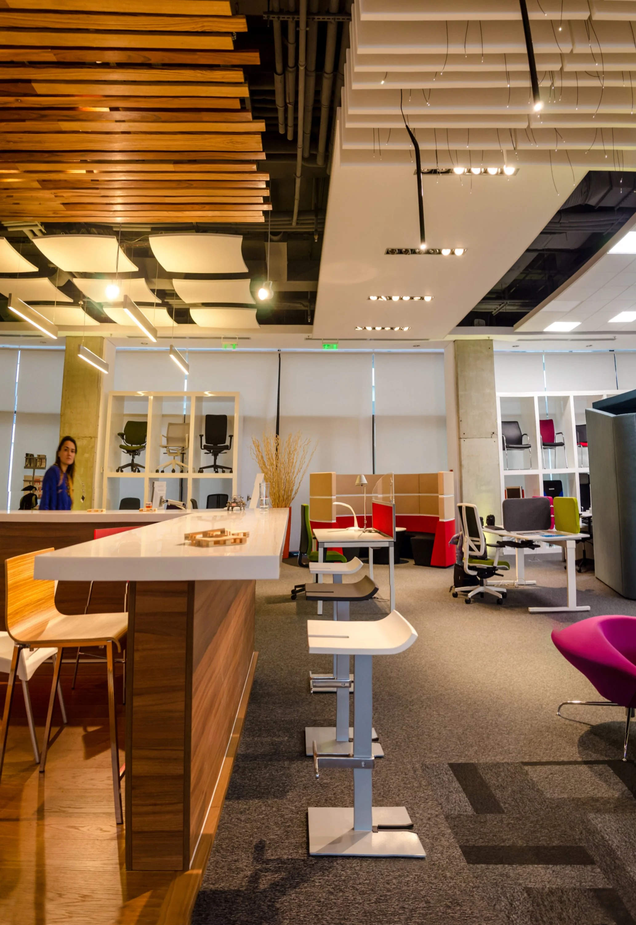 Modern office space with a wooden counter at the front, open workspace with desks and colorful chairs, shelves displaying office chairs, ceiling with decorative panels and lighting fixtures, and a woman standing near the counter.