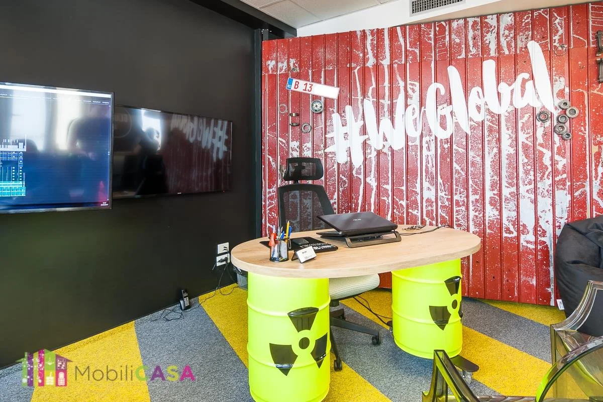 An office with a black wall on the left, two large flat-screen monitors, and a corner wall with red wood paneling and a white hashtag sign reading "#HogWild". The desk is made from a wooden tabletop supported by two yellow barrels with black radiation symbols, and has office supplies and a laptop on it.