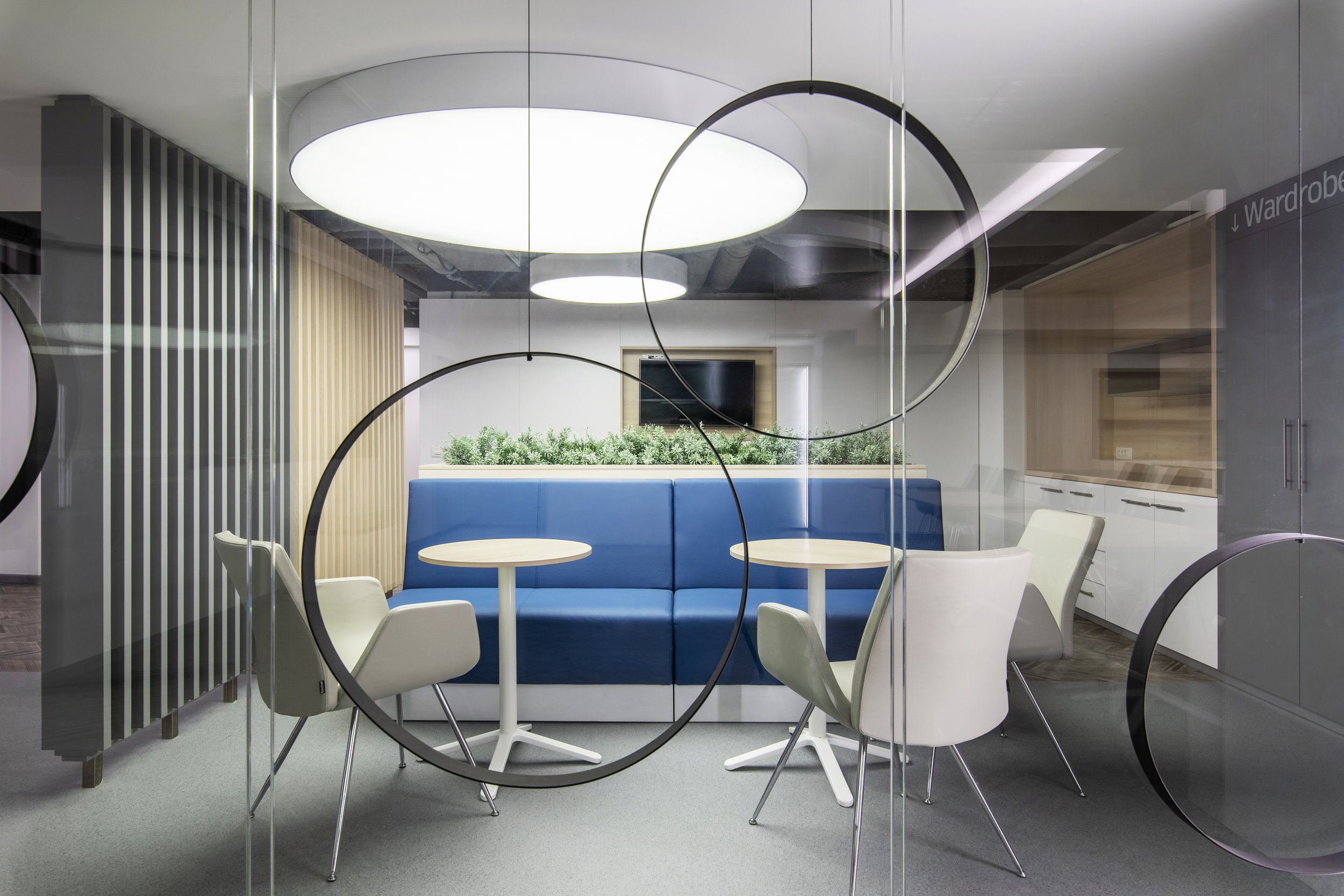 Modern office lounge with blue upholstered bench, white chairs, round white tables, and circular black decor elements on glass wall, featuring ceiling lights, a TV on the wall, and green plants in the background.