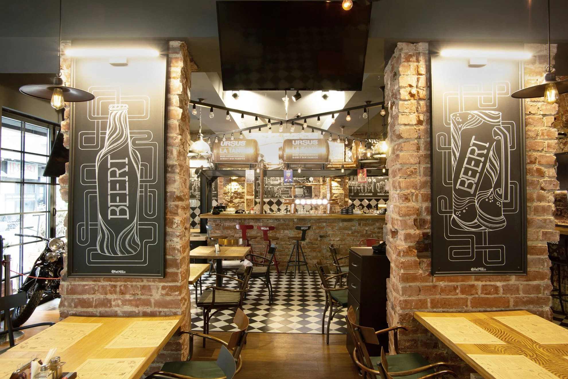 Interior of a restaurant or pub with exposed brick walls, black and white checkered flooring, wooden tables, and chairs. There are large blackboards with beer-themed chalk art, string lights, and a bar area in the background.