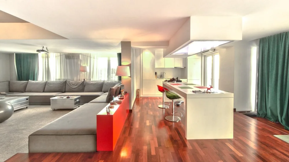 Open-concept living room and kitchen with hardwood floors, L-shaped gray sofa, red accent wall, white kitchen island with colorful bar stools, and large windows with green curtains.
