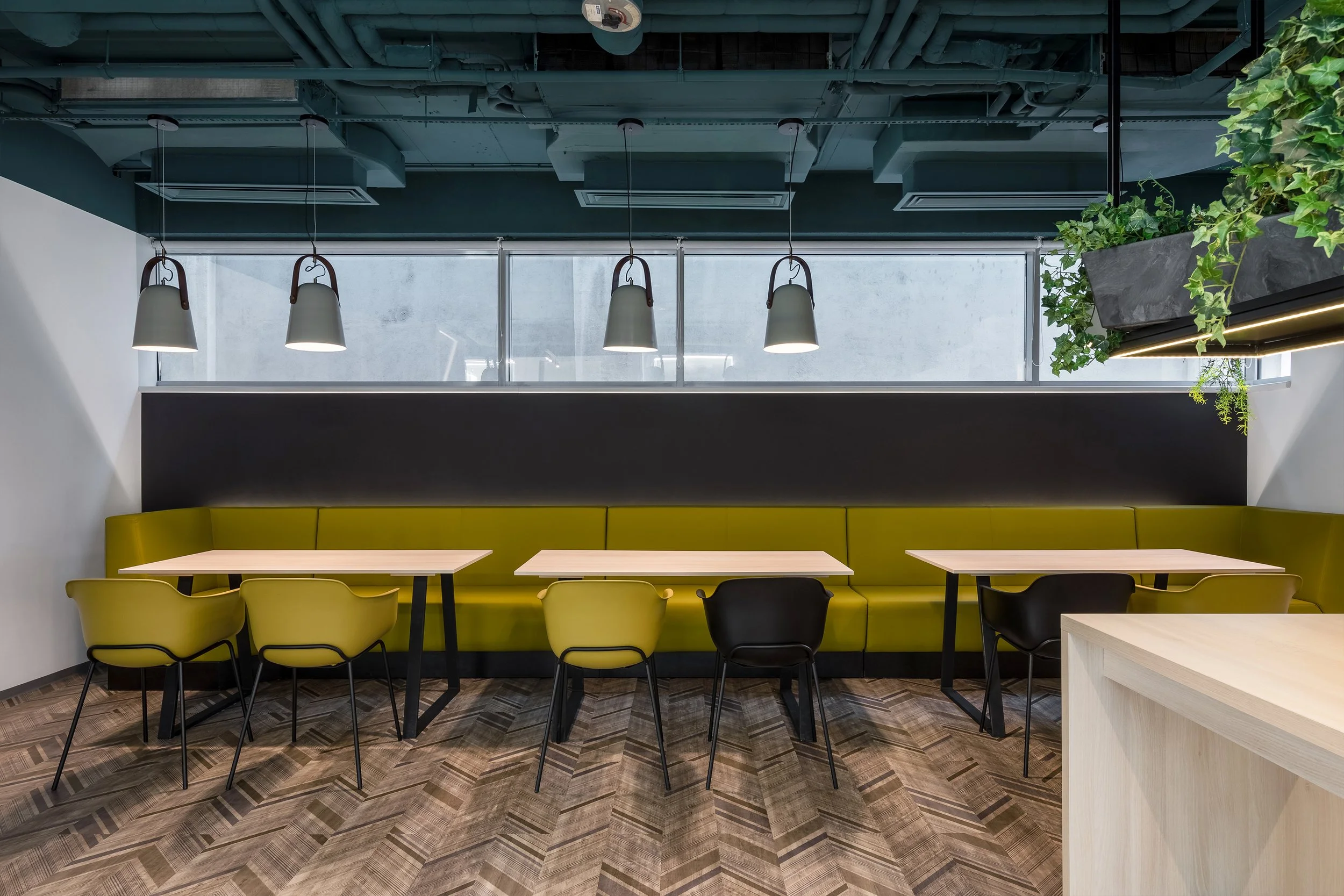 Modern restaurant seating area with yellow booths and chairs, a black wall behind the booths, hanging pendant lights, and a window with a concrete building outside. Green plants are also visible.