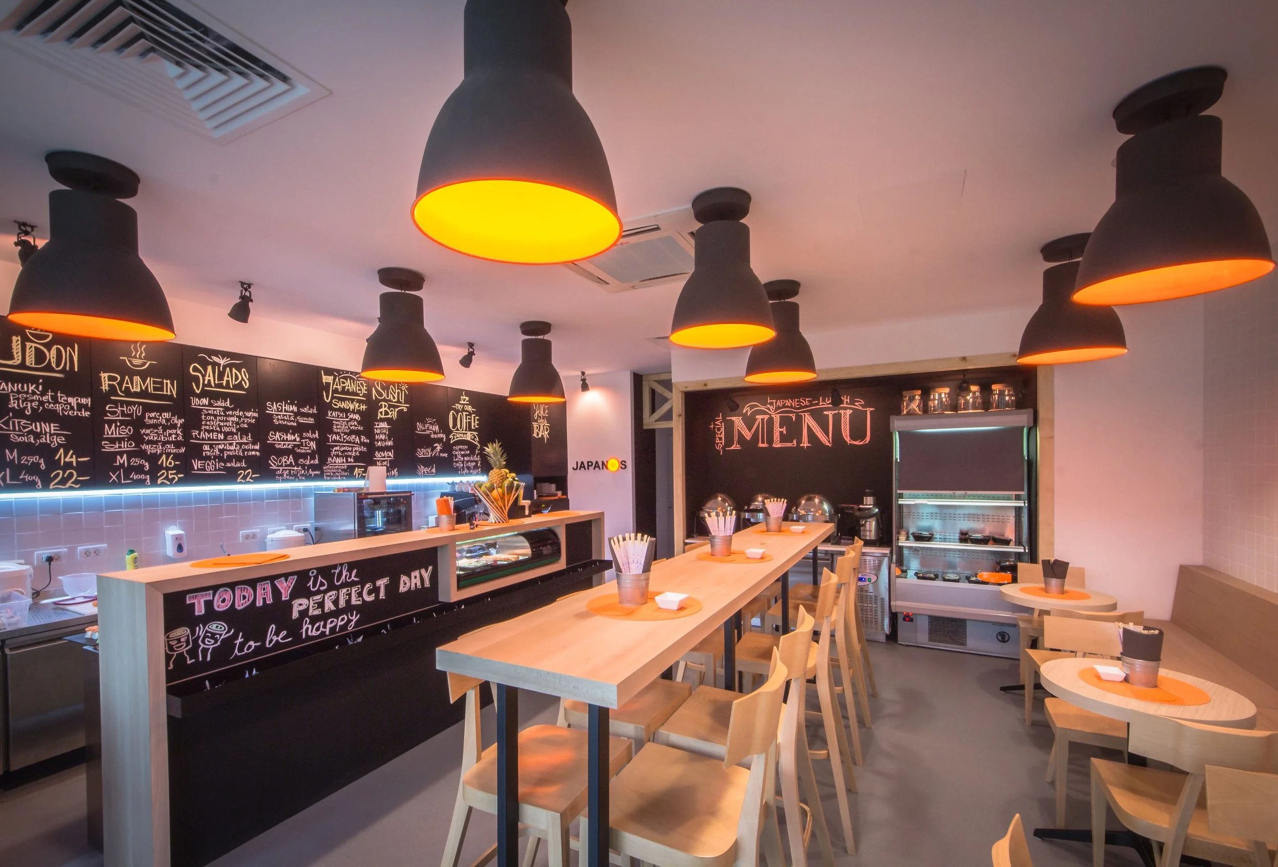 Interior of a modern Japanese restaurant with black pendant lights, a chalkboard menu, and a table set with orange placemats, napkins, and utensils.
