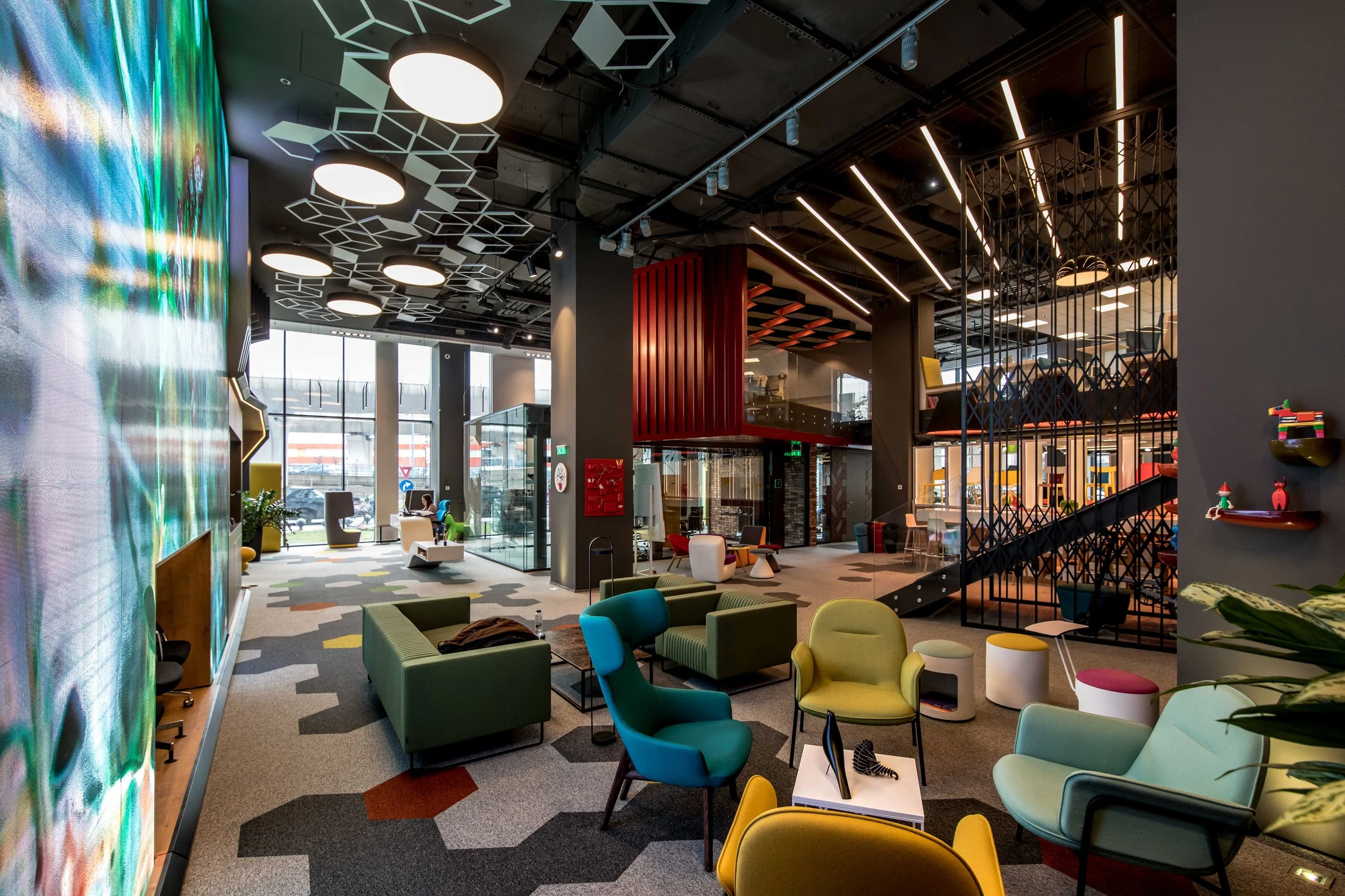 Modern lobby or lounge area with colorful chairs, sofas, and stools, large windows, patterned carpet, and high ceilings with geometric and linear lighting fixtures.