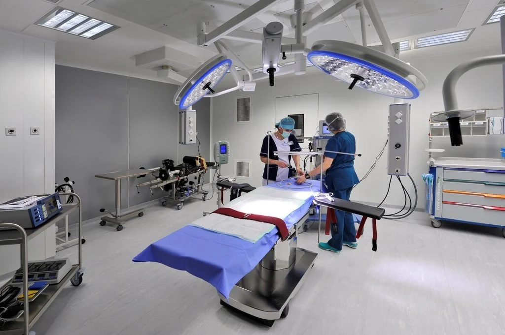 An operating room with two medical professionals preparing for surgery, featuring surgical lights, equipment, and an operating table in the center.