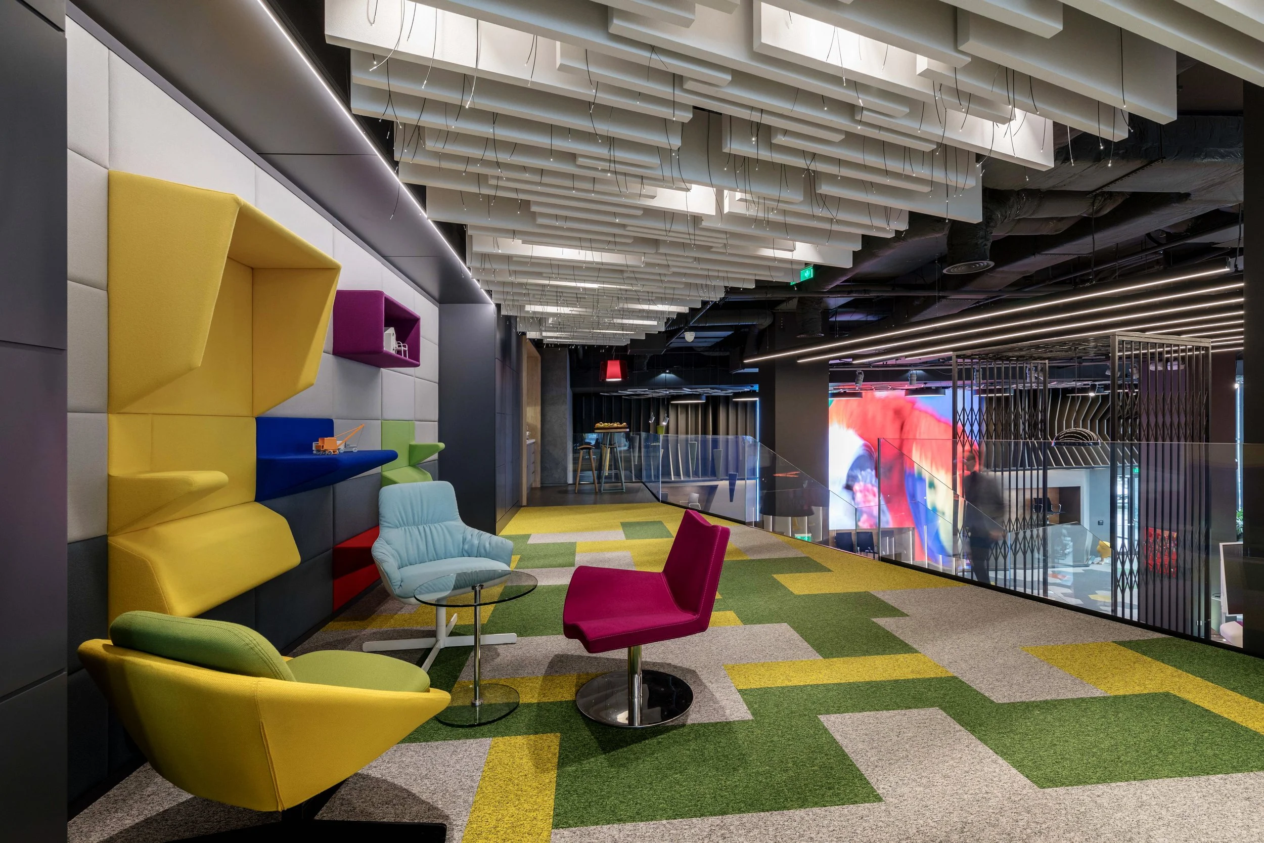 Modern, colorful lounge area with abstract patterned carpet, stylish chairs, and decorative wall panels in an indoor space with ceiling installations and large screen display in the background.