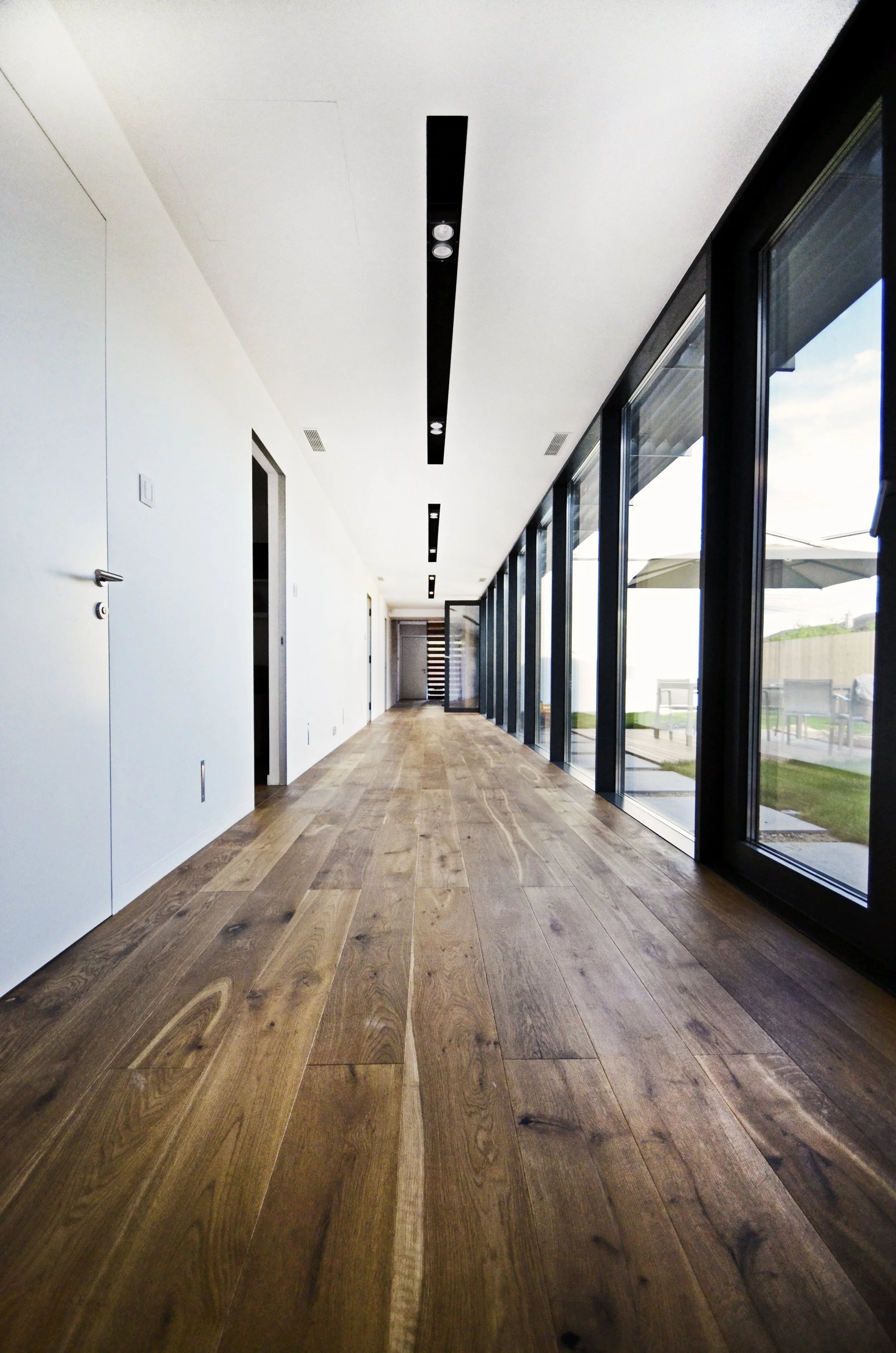 Modern interior hallway with wooden flooring, white walls, and large glass windows showing outdoor patio area.