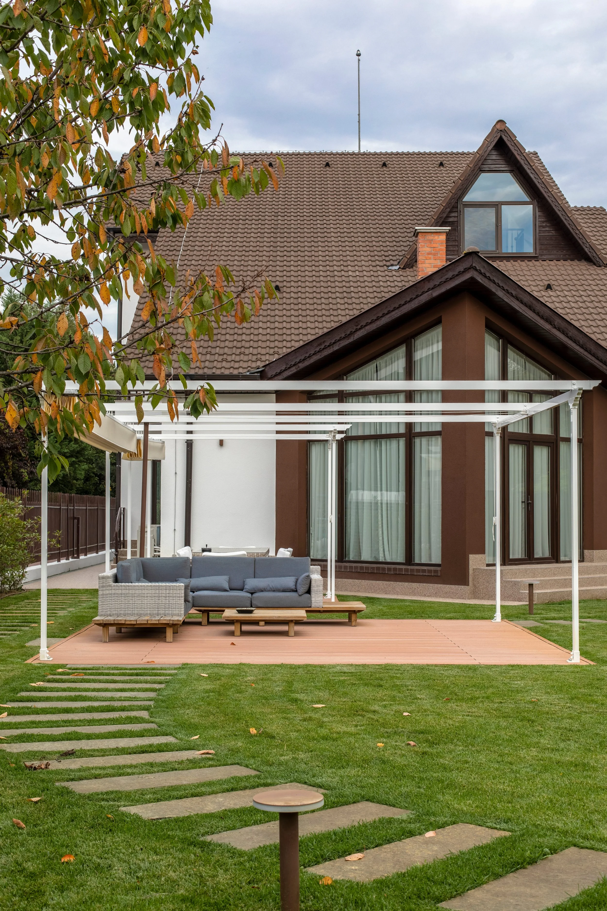 A backyard patio with a gray outdoor sofa set on a wooden deck, a white trellis, green grass with stepping stones, and a large modern house with glass windows and brown walls in the background. A tree with green and orange leaves is in the foreground.