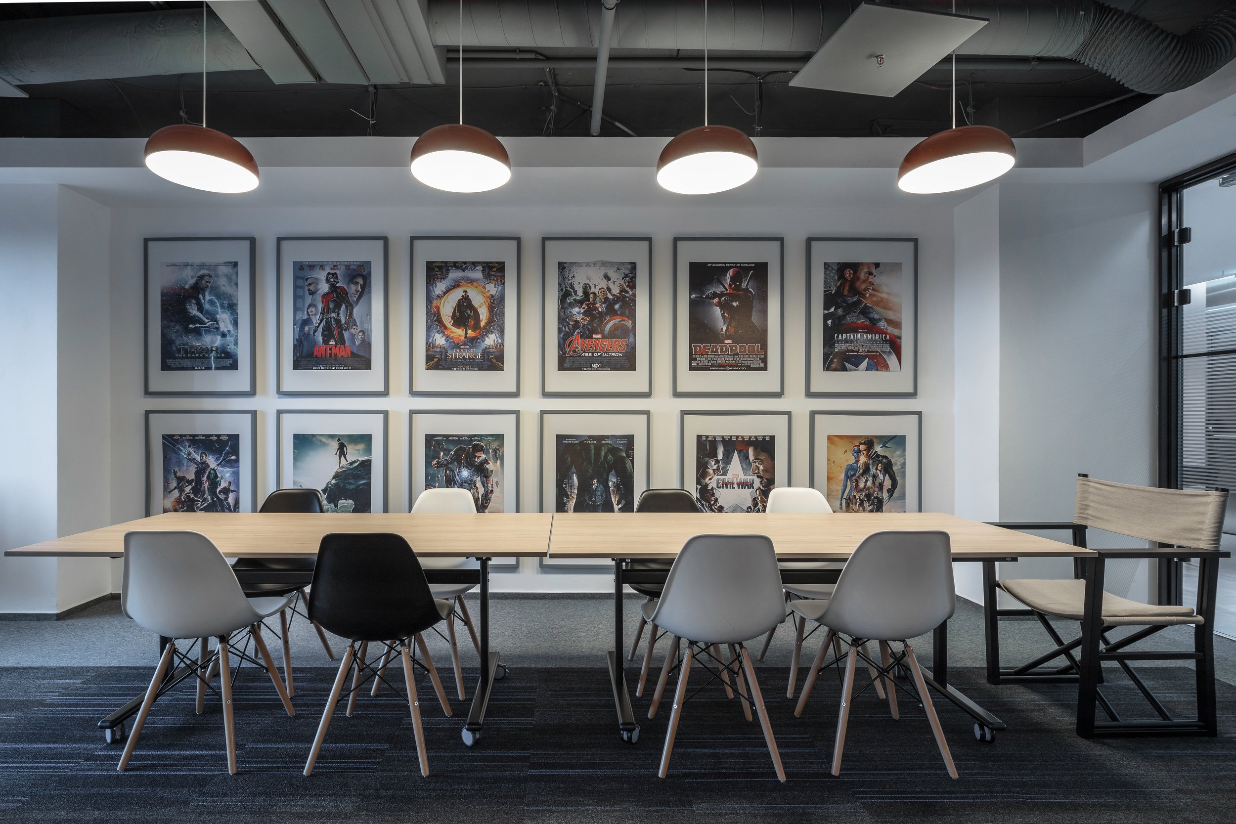 Conference room with a long wooden table, white, black, and beige chairs, framed movie posters on the wall, and modern lighting fixtures overhead.