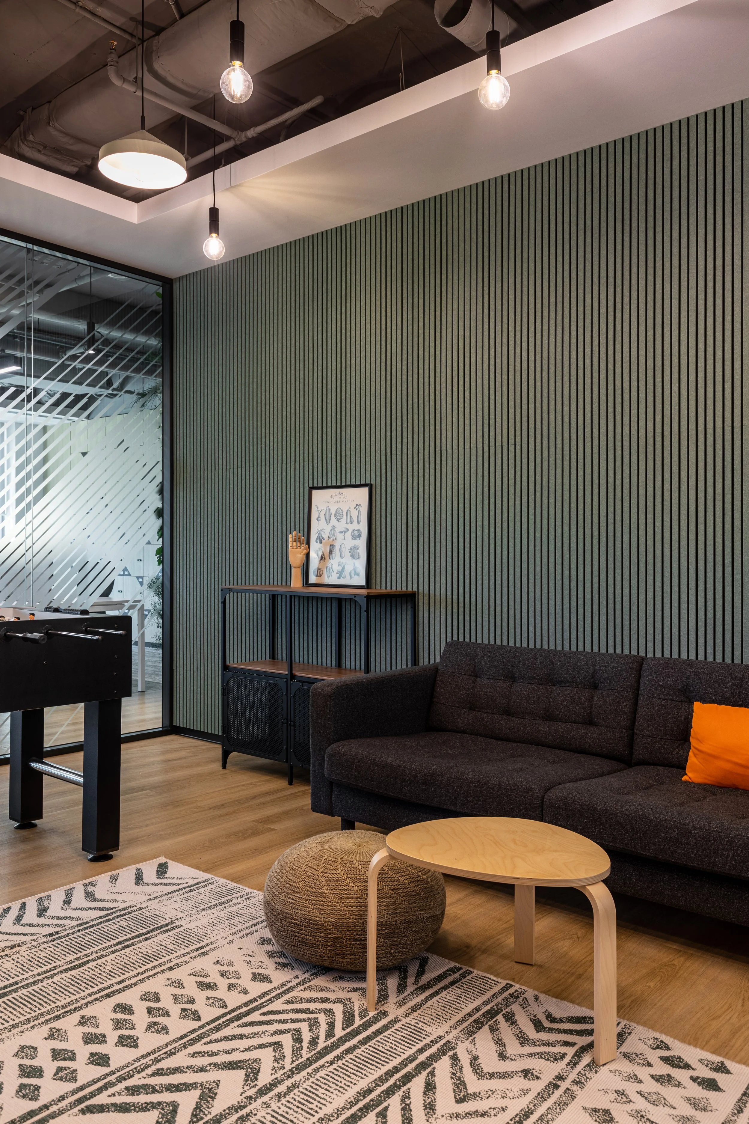 Modern living room with black sofa, wooden coffee table, textured pouf, patterned rug, green striped accent wall, and glass partition.