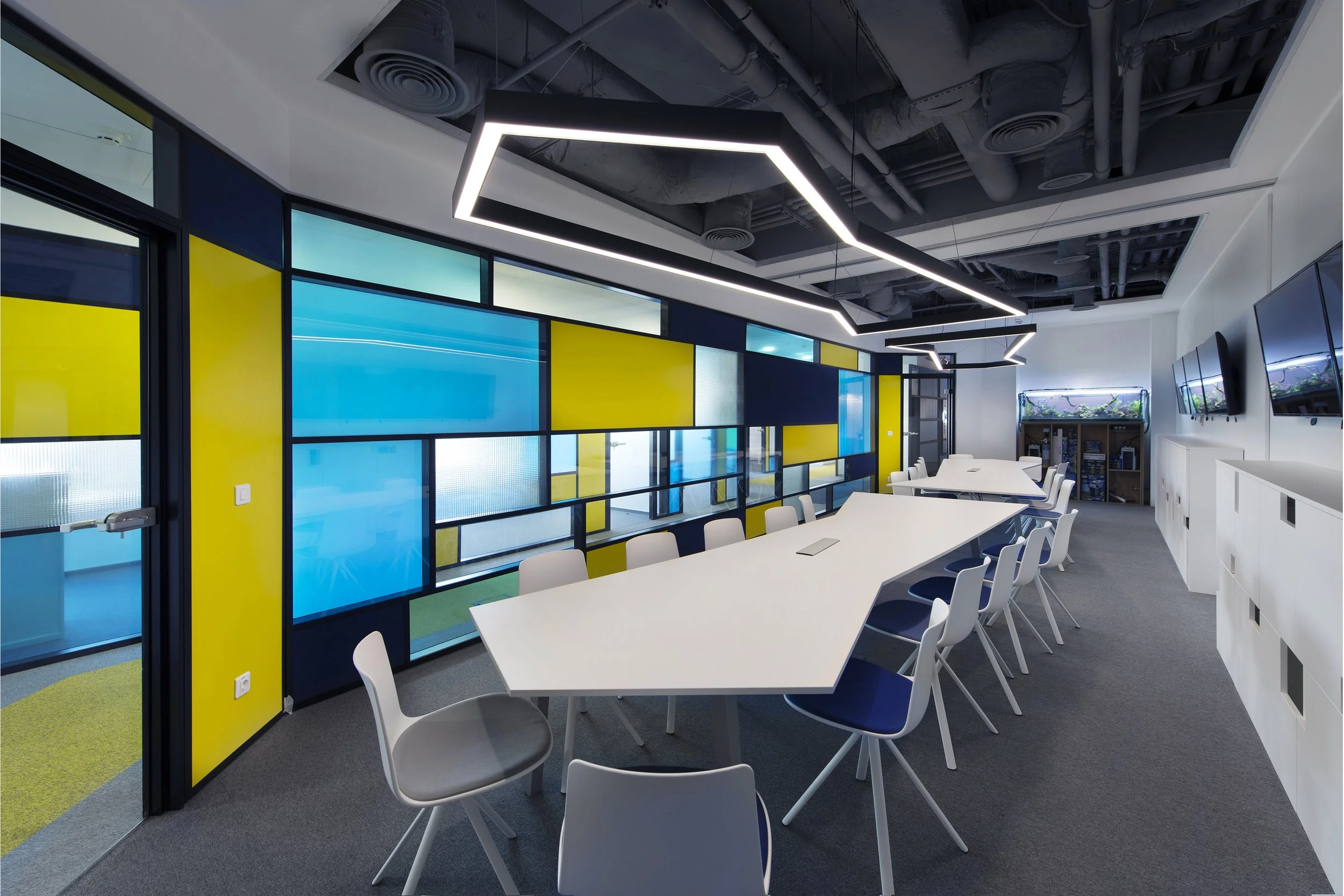 Modern conference room with a long white table, white and black chairs, multiple wall-mounted flat-screen TVs, a geometric ceiling light fixture, and a colorful glass wall with blue, yellow, and clear panels.
