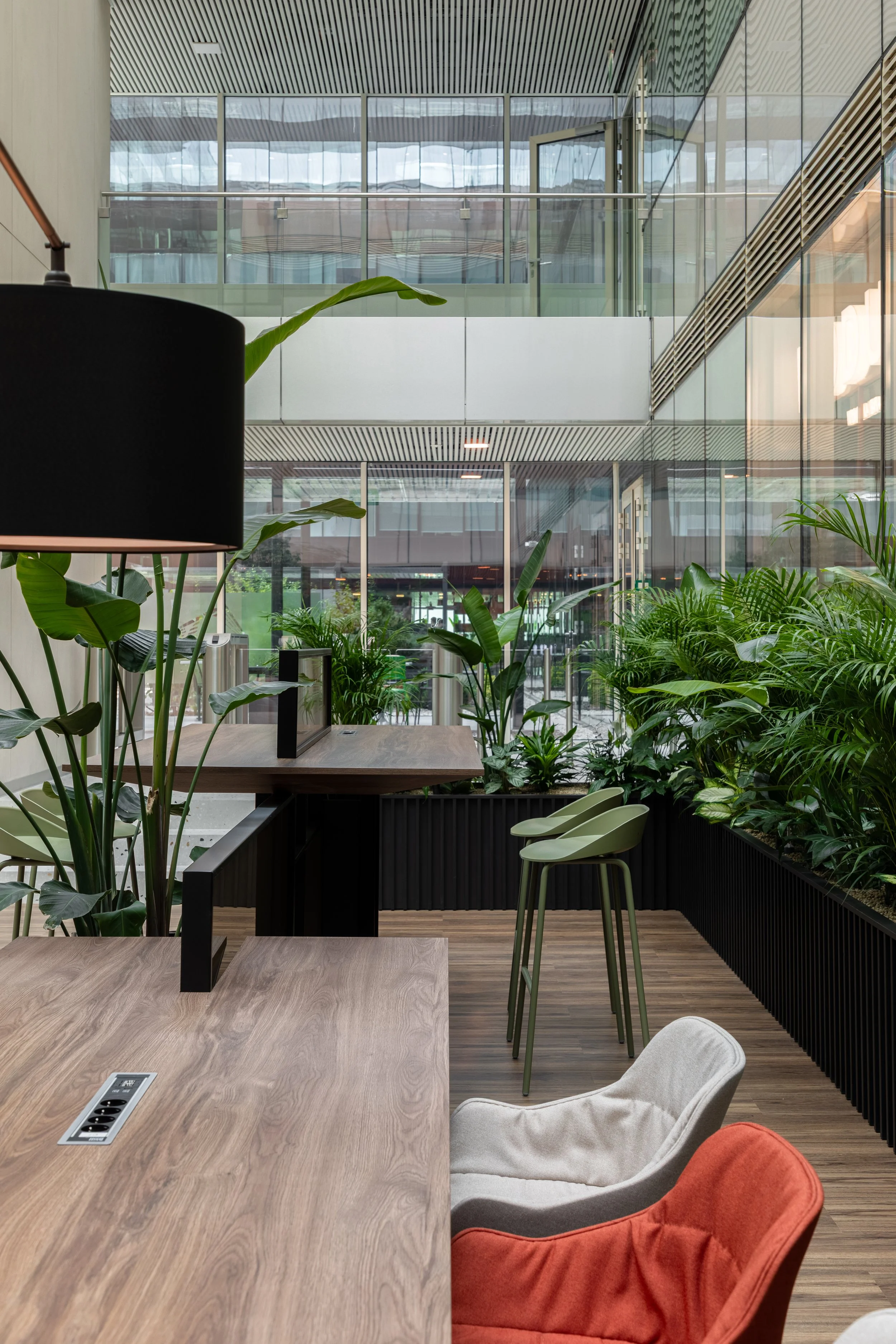 Modern indoor space with wooden tables, green chairs, and lush green plants, with large glass windows and a multi-level layout.