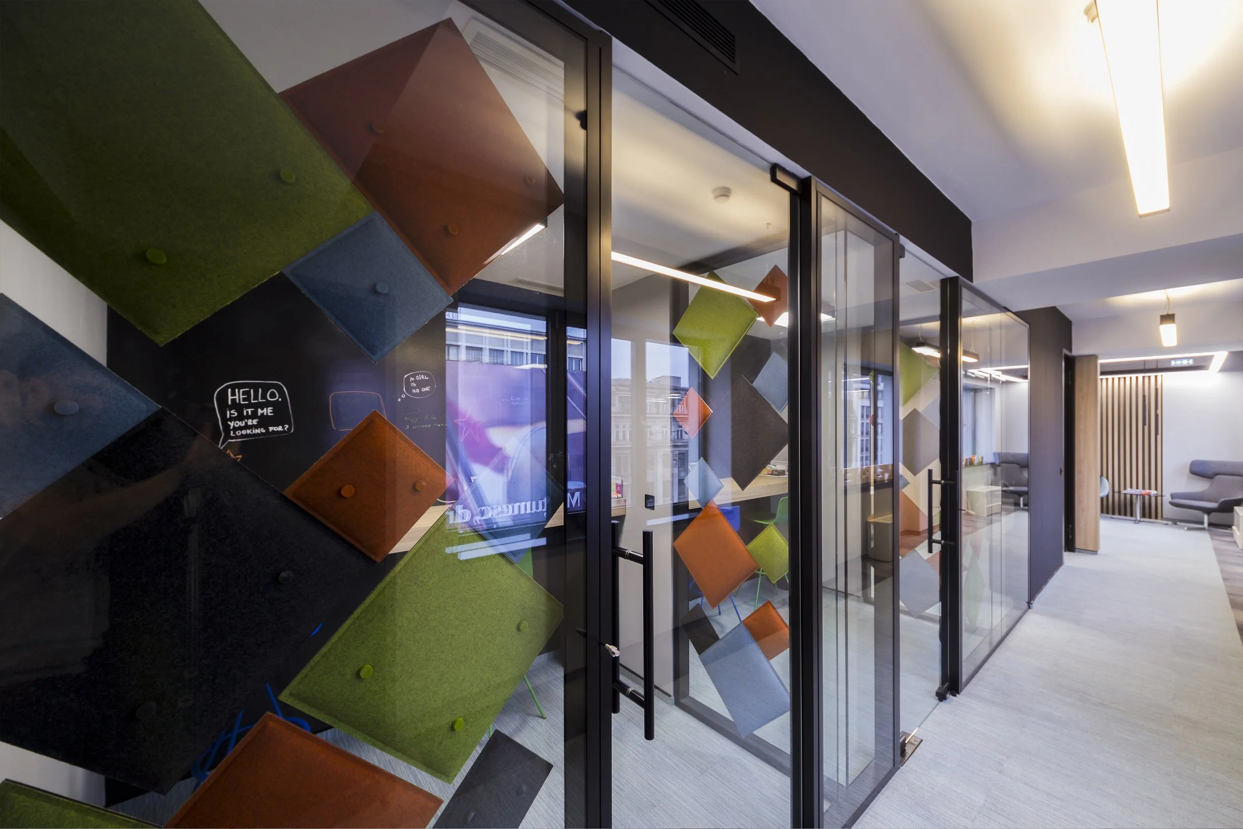 Modern office interior with colorful acoustic panels on glass partitions and seating area with chairs and sofas.