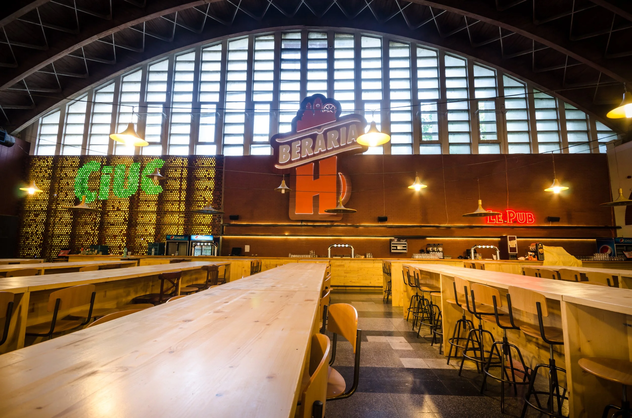 Interior of a rustic pub with wooden tables and chairs, large windows, and colorful neon signs that say 'Crush', 'Beraria', and 'Le. Pub'.