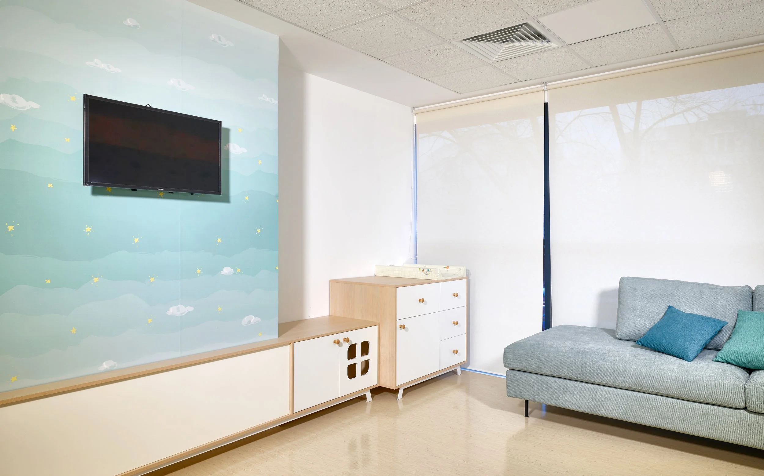 A modern living room with a light gray couch, blue and teal throw pillows, a white and wood sideboard, and a flat screen TV on a pastel cloud-themed wall. Sunlight filters through a partially covered window with a view of leafless trees outside.