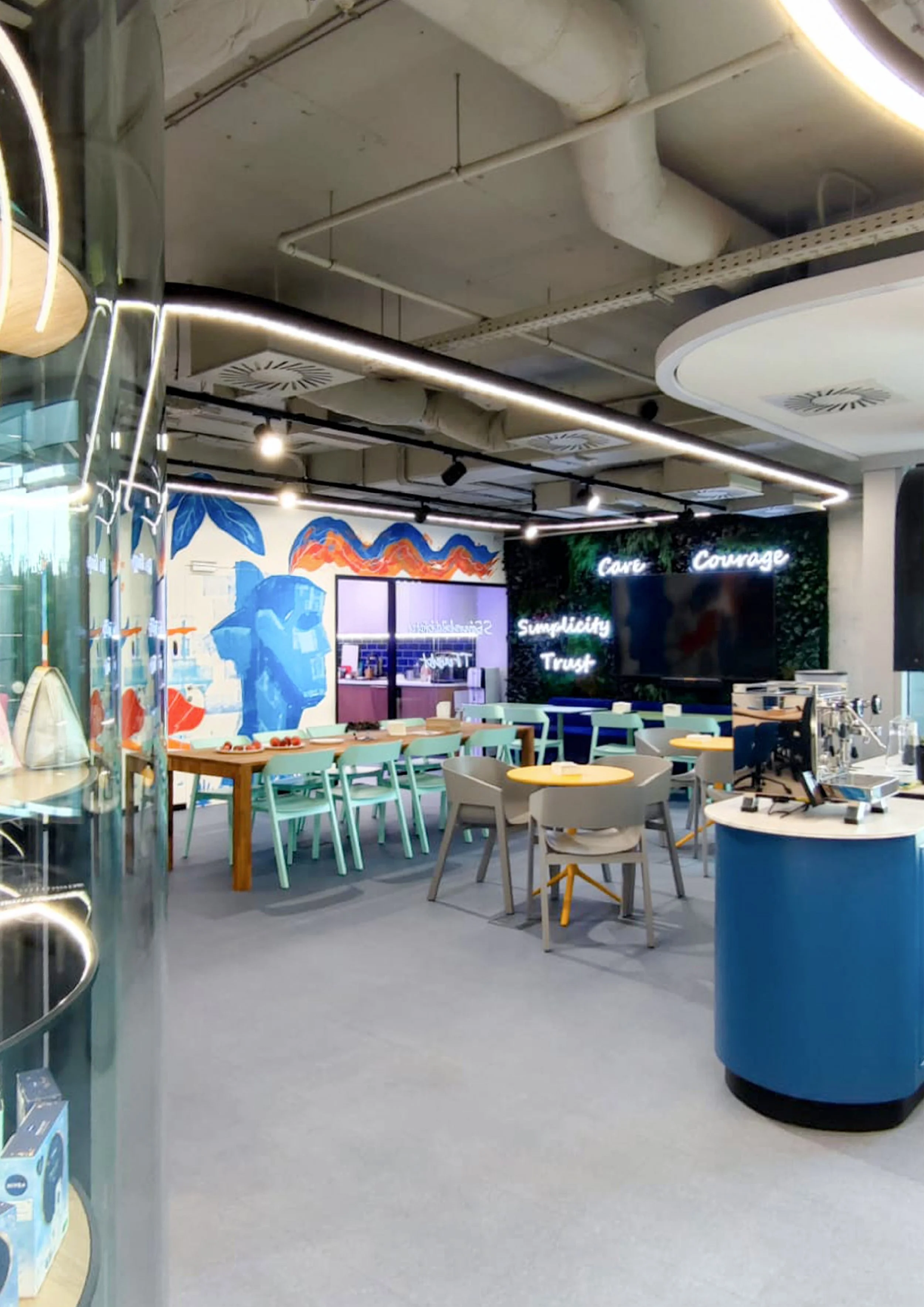 This image shows a modern, brightly lit cafe with a colorful mural on the wall, neon signs with words like 'Cure,' 'Courage,' 'Simplicity,' and 'Trust,' and a mix of tables and chairs for seating. There is also a blue counter with coffee equipment.
