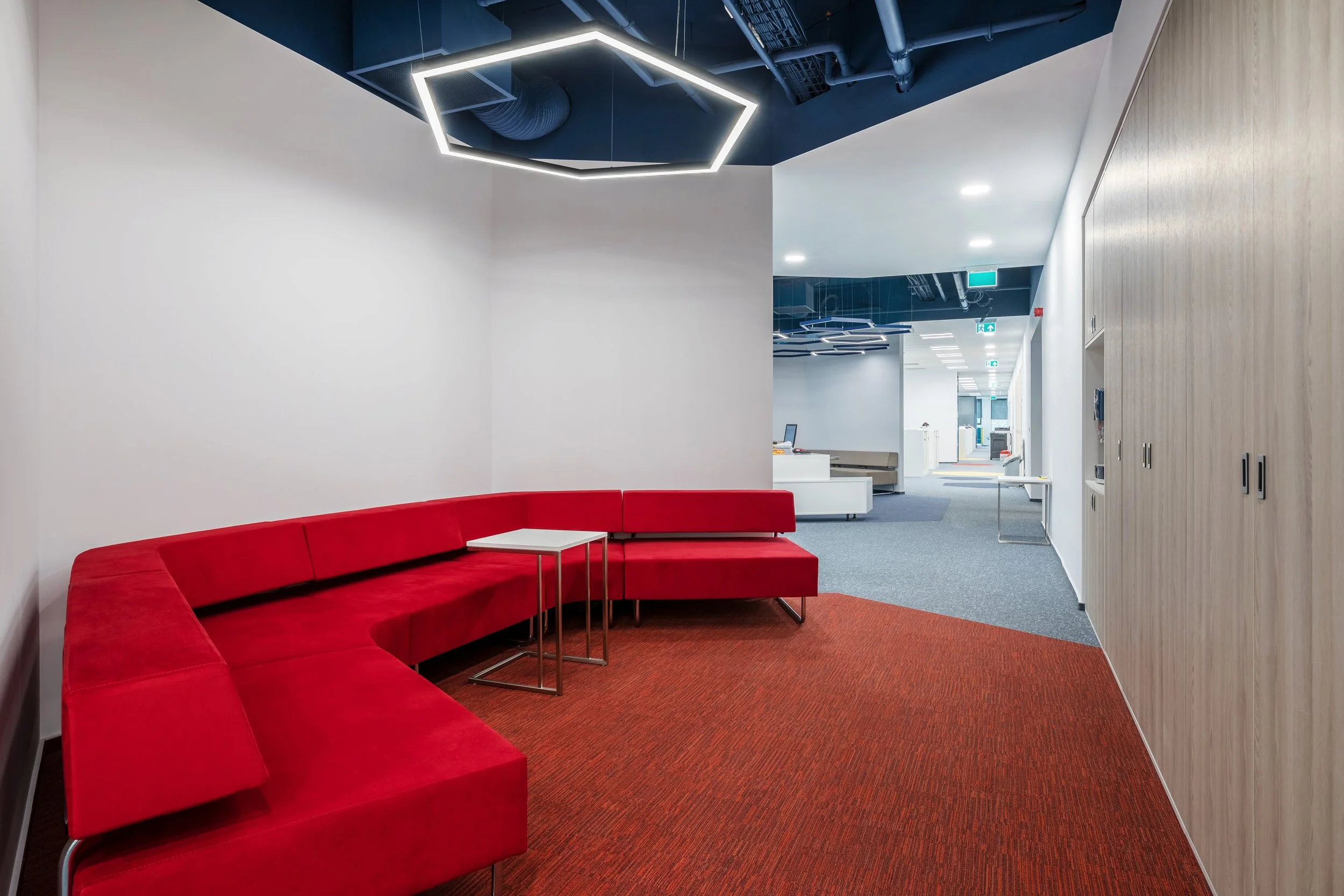 Modern office lounge with red sectional sofa, small white table, and a mix of carpeted and tiled floors, illuminated by a hexagonal LED ceiling light.