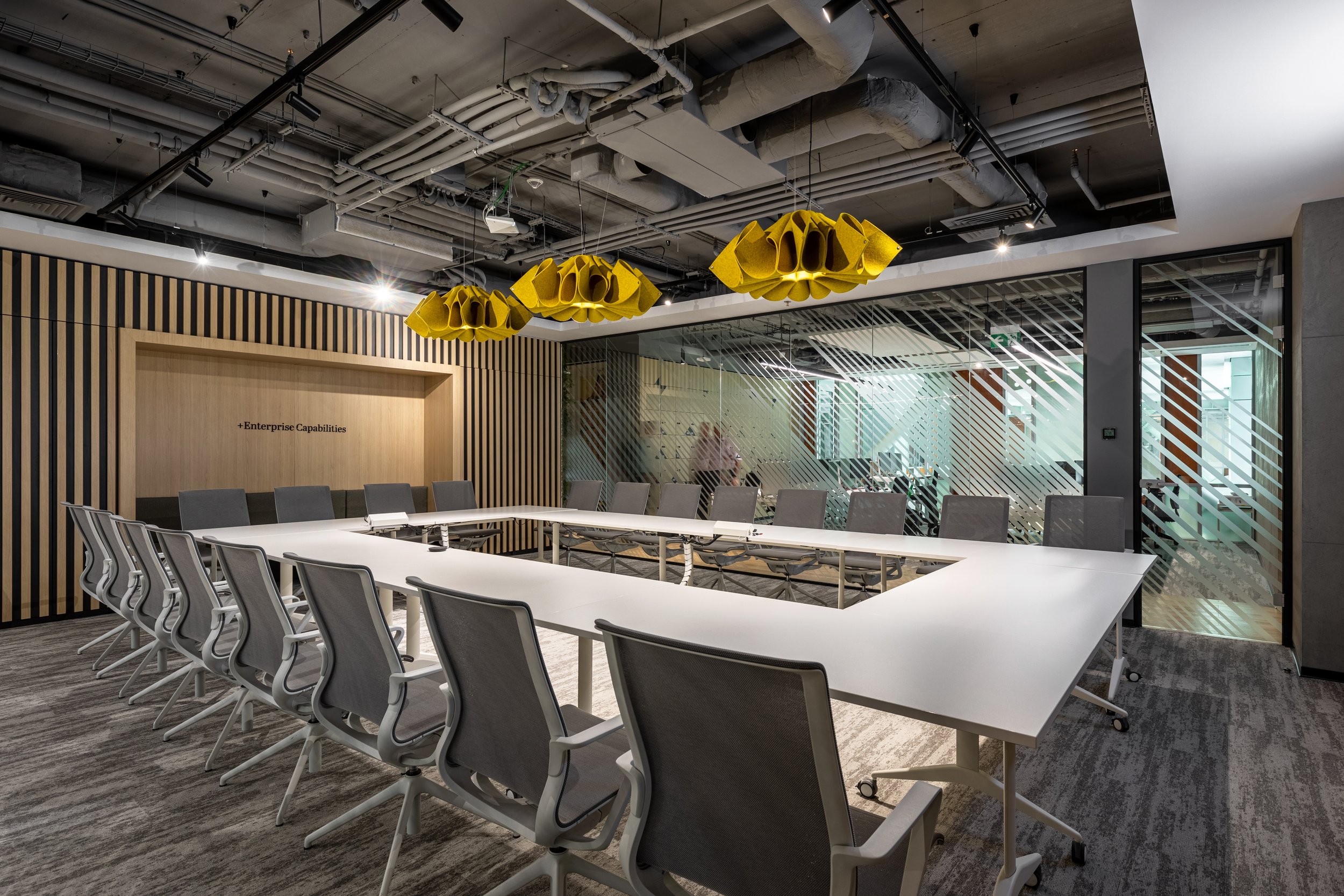 Modern conference room with a U-shaped white table, gray office chairs, a wooden wall with vertical black slats, and three yellow sculptural light fixtures hanging from the ceiling. There are glass walls with diagonal striped patterns and an exposed ceiling with visible pipes and ducts.