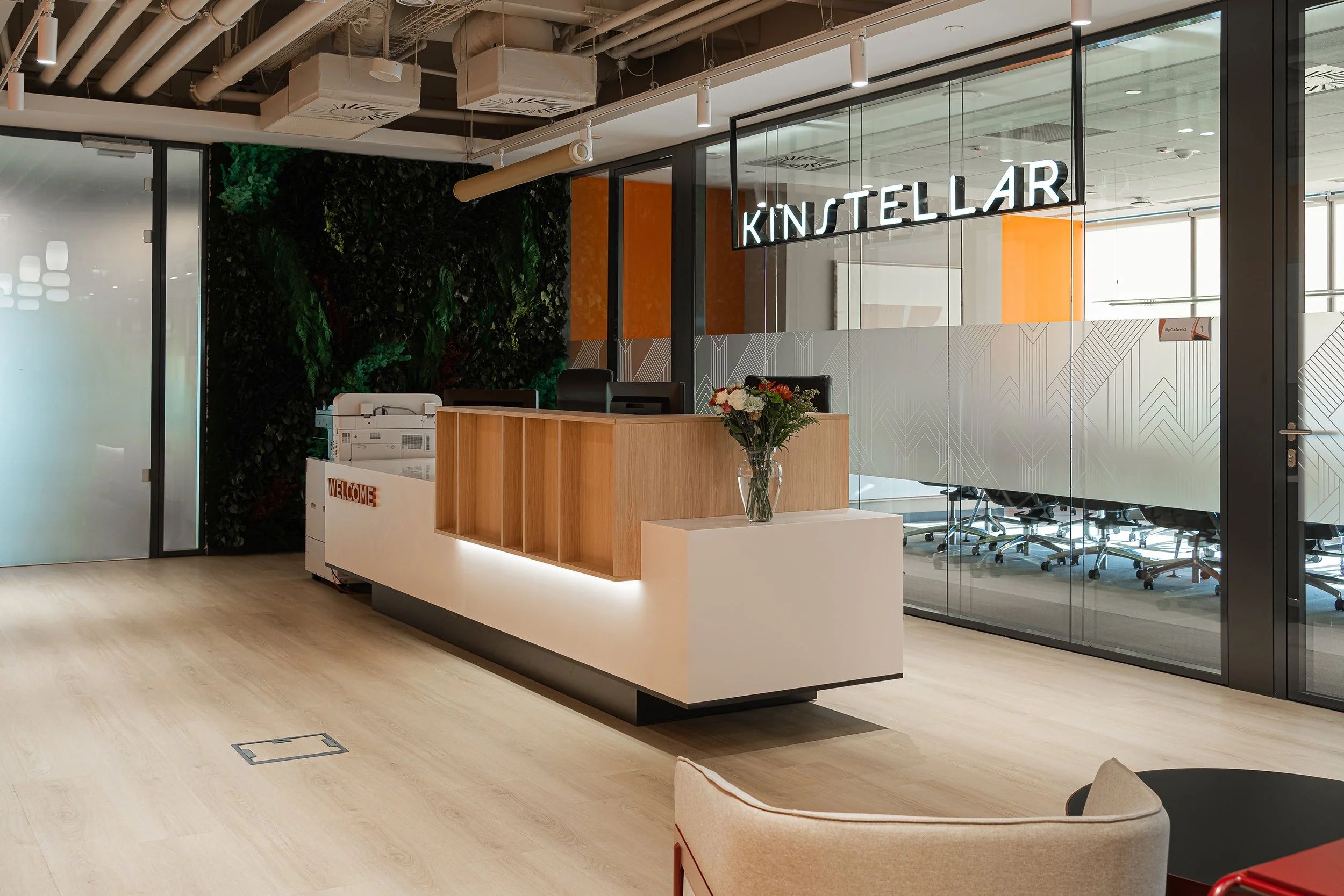 Modern office reception area with a wooden and white front desk, a vase of flowers, a green wall, glass walls.