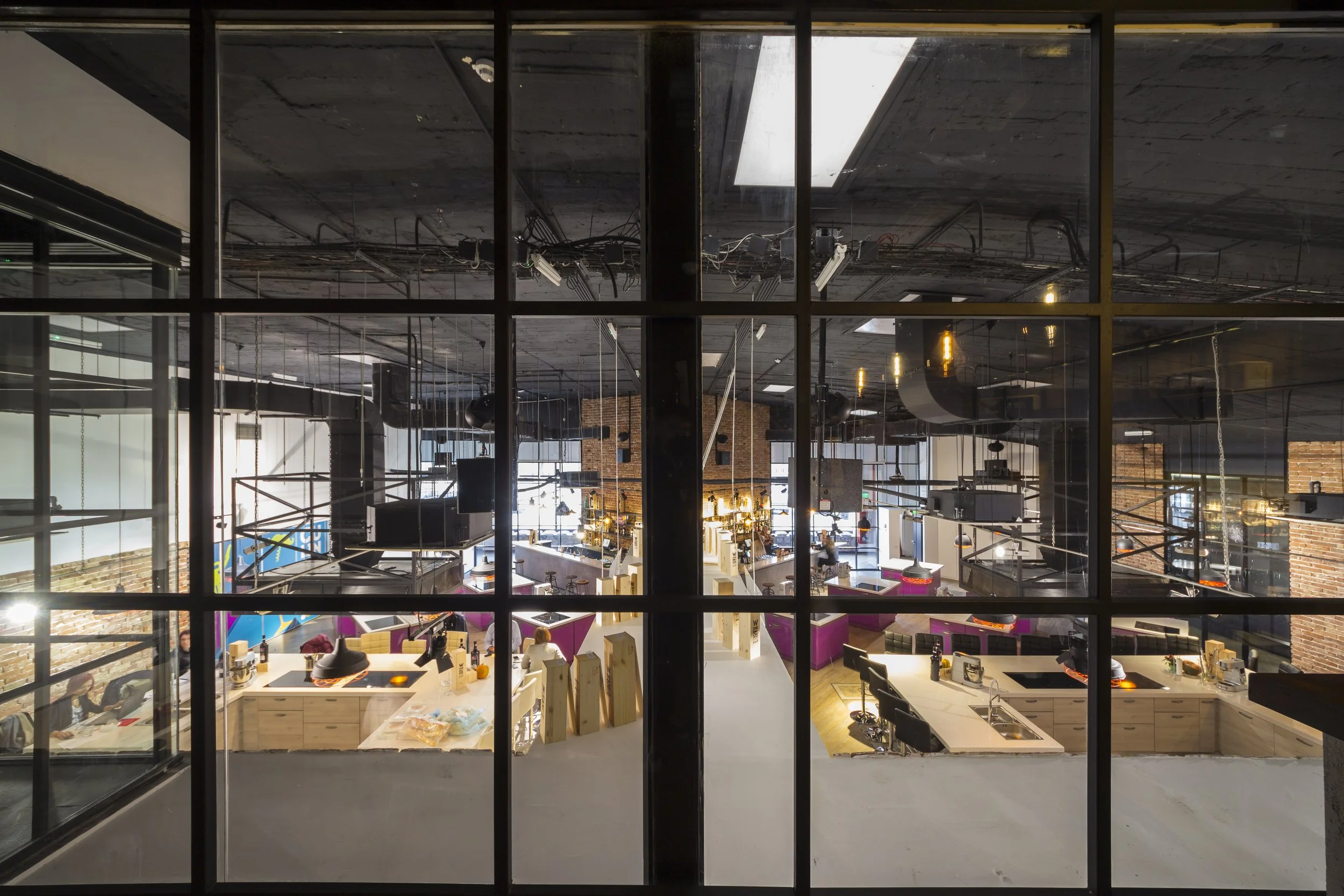 Interior view of a modern office space with open layout, workstations, and exposed ceiling pipes as seen through large window panes.