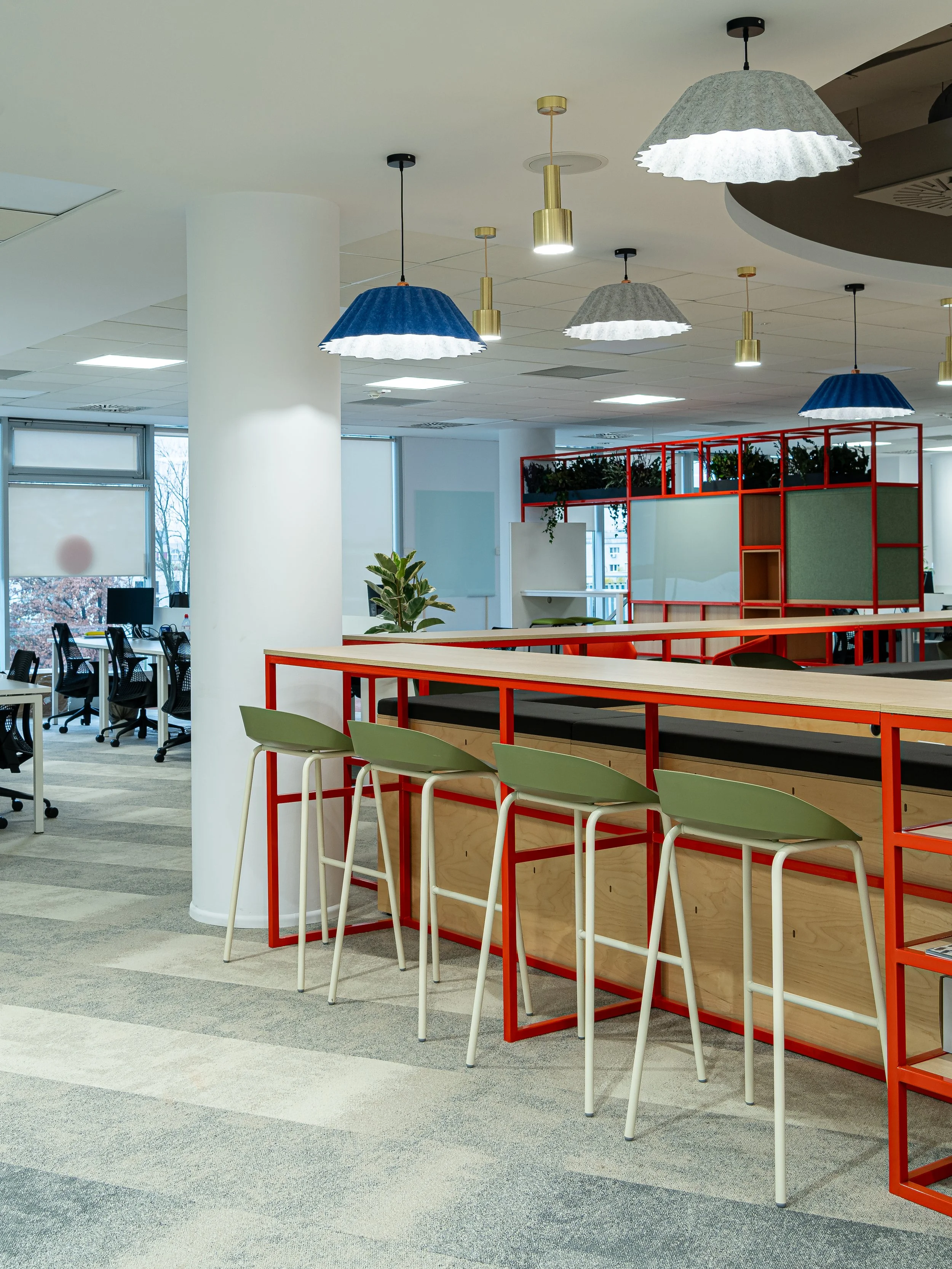Interior of a modern office with bar-height chairs, a high counter with a wooden top and red metal frame, hanging pendant lights in blue, gray, and white, and office desks with chairs near windows.