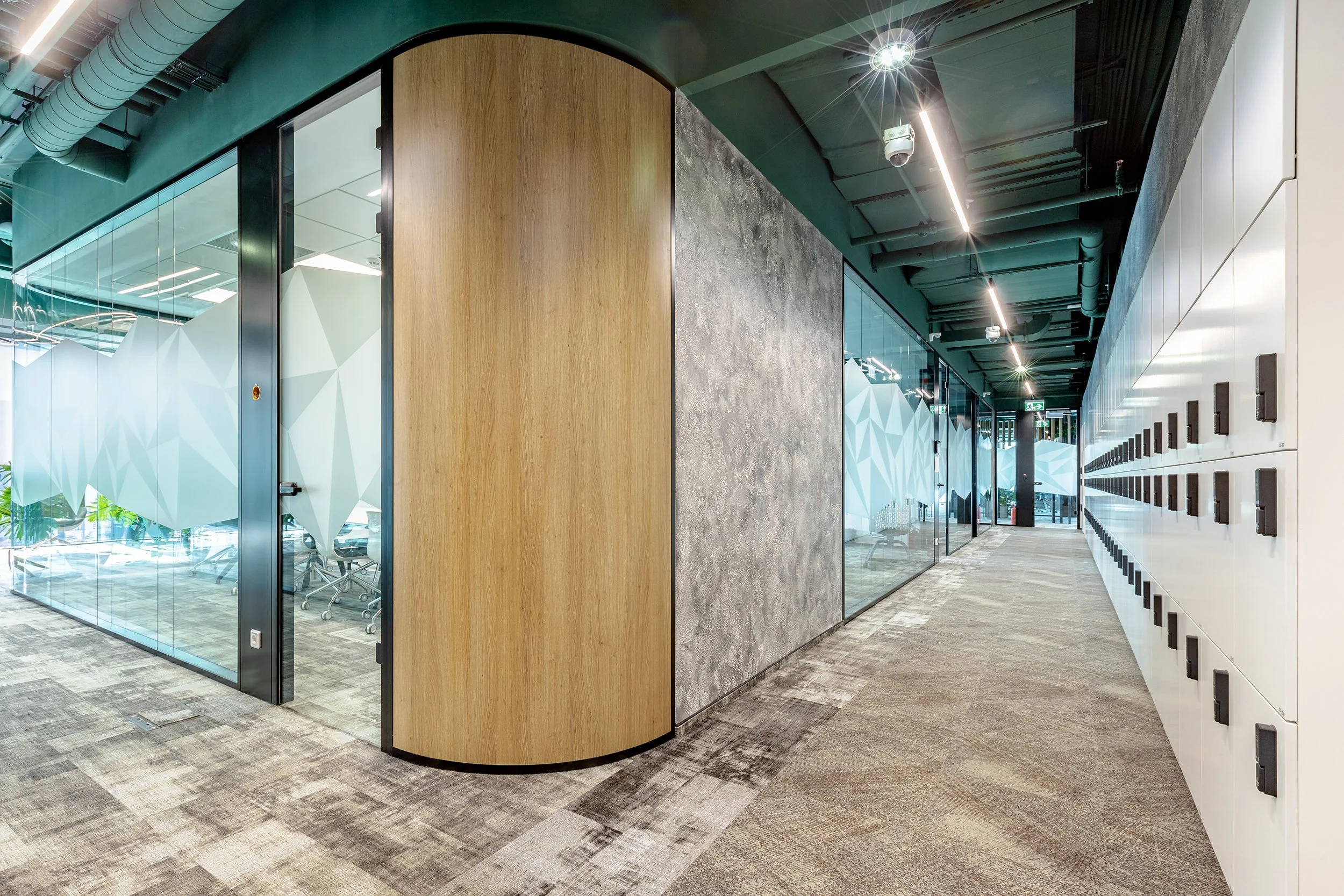 Modern office corridor with glass-walled conference rooms, wood and concrete walls, gray carpeted floor, row of white lockers with black locks, industrial ceiling with exposed ducts and slim lighting.