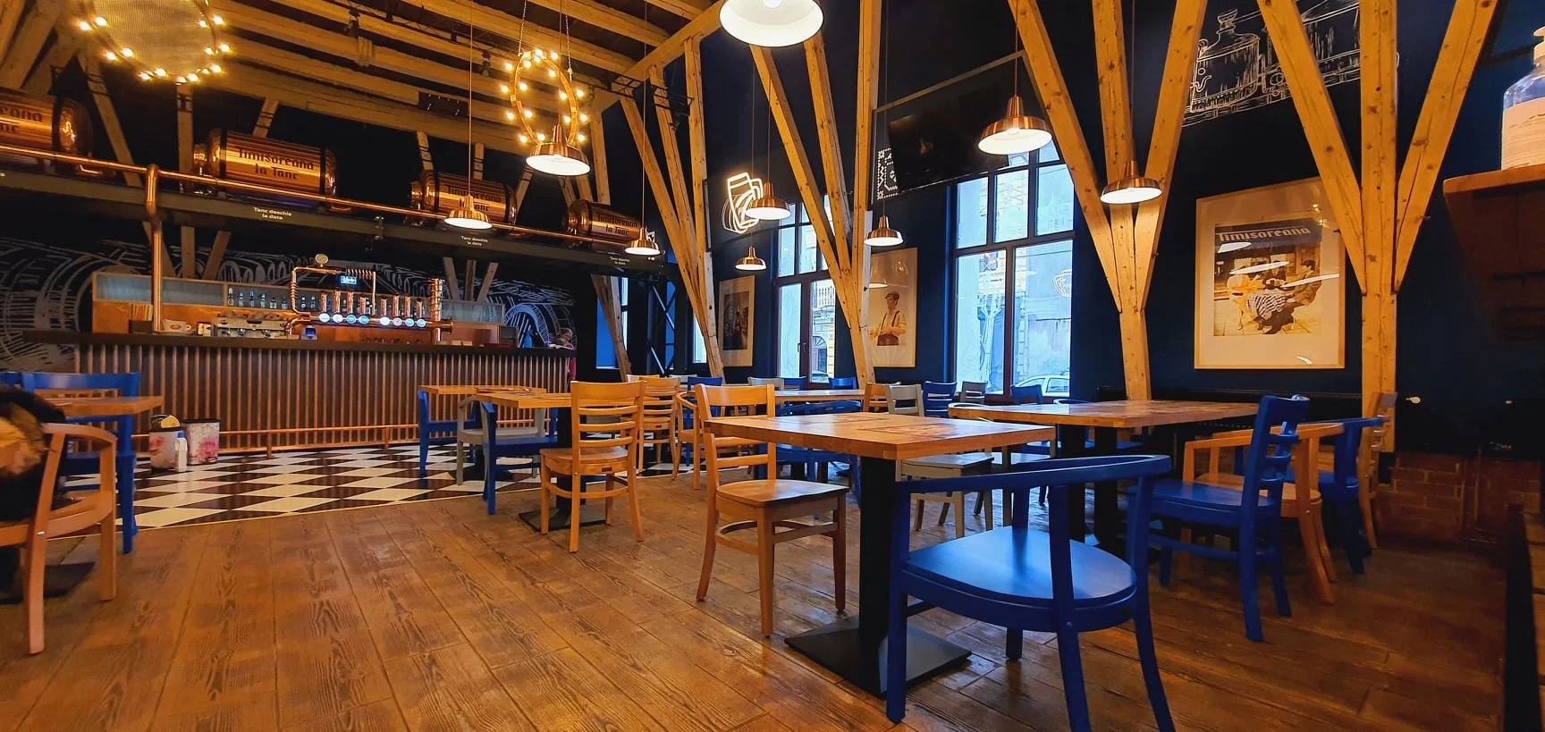 Interior of a stylish restaurant with wooden furniture, high ceilings with exposed beams, large windows, wall art, and warm ambient lighting.