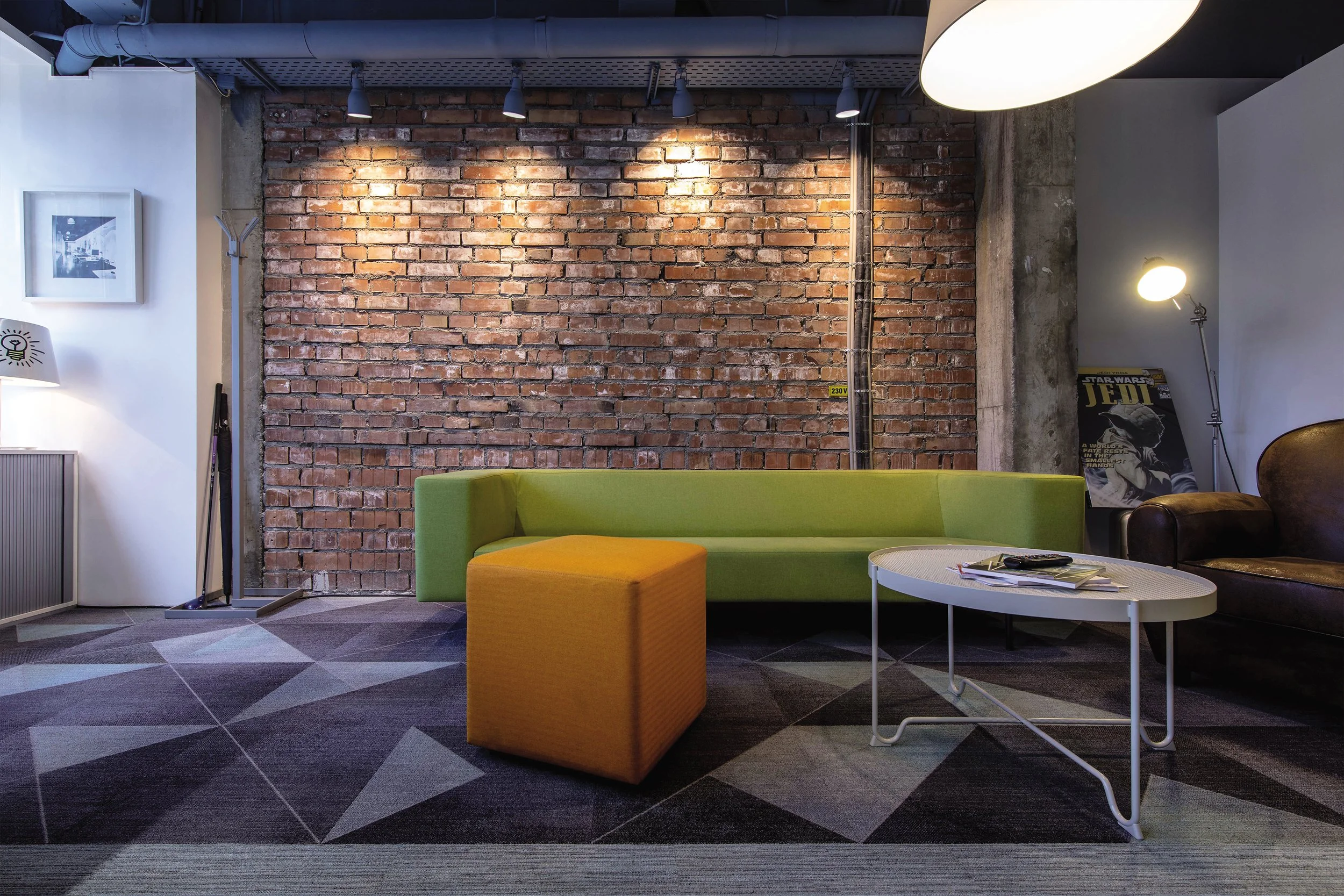 A modern lounge area with a green sofa, yellow ottoman, round white coffee table, and brown leather armchair set against a brick wall with ceiling spotlights and a standing lamp. A Star Wars poster is leaning against the wall.