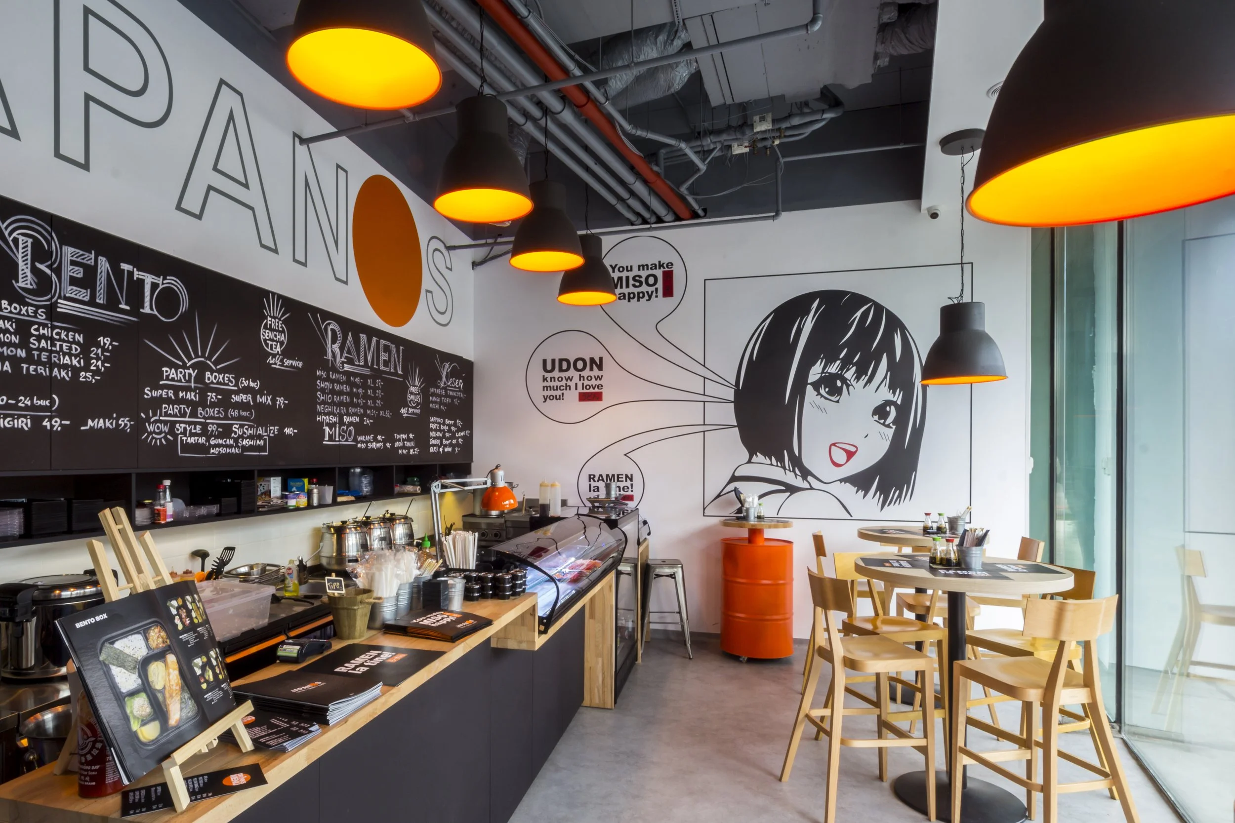 Interior of a modern Japanese restaurant featuring a black chalkboard menu, a large mural of a stylized anime girl, and dining tables with chairs, with orange hanging pendant lights.