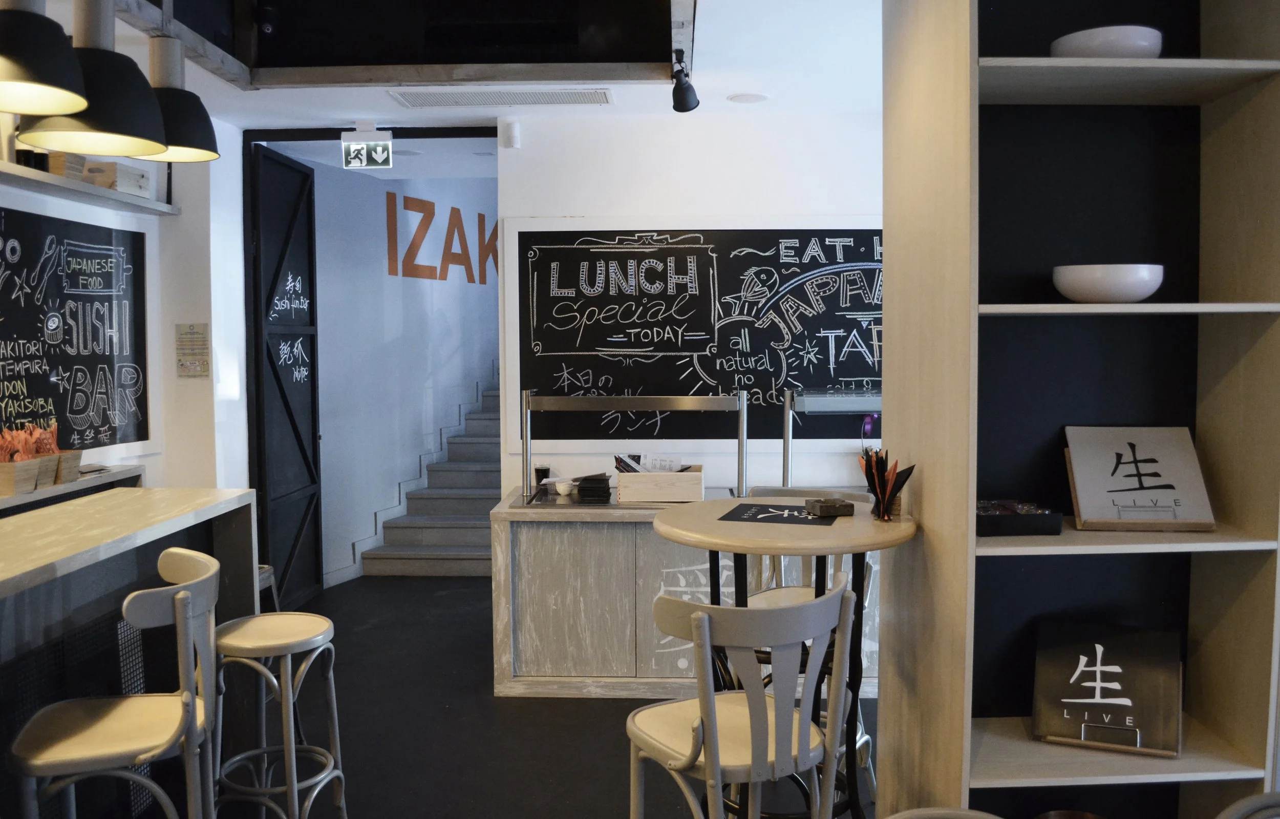 Interior of a modern restaurant with black chalkboard wall featuring handwritten menu items, light-colored chairs, a small round table with black menu and utensils, black and white shelving with decorative items, and a staircase in the background.