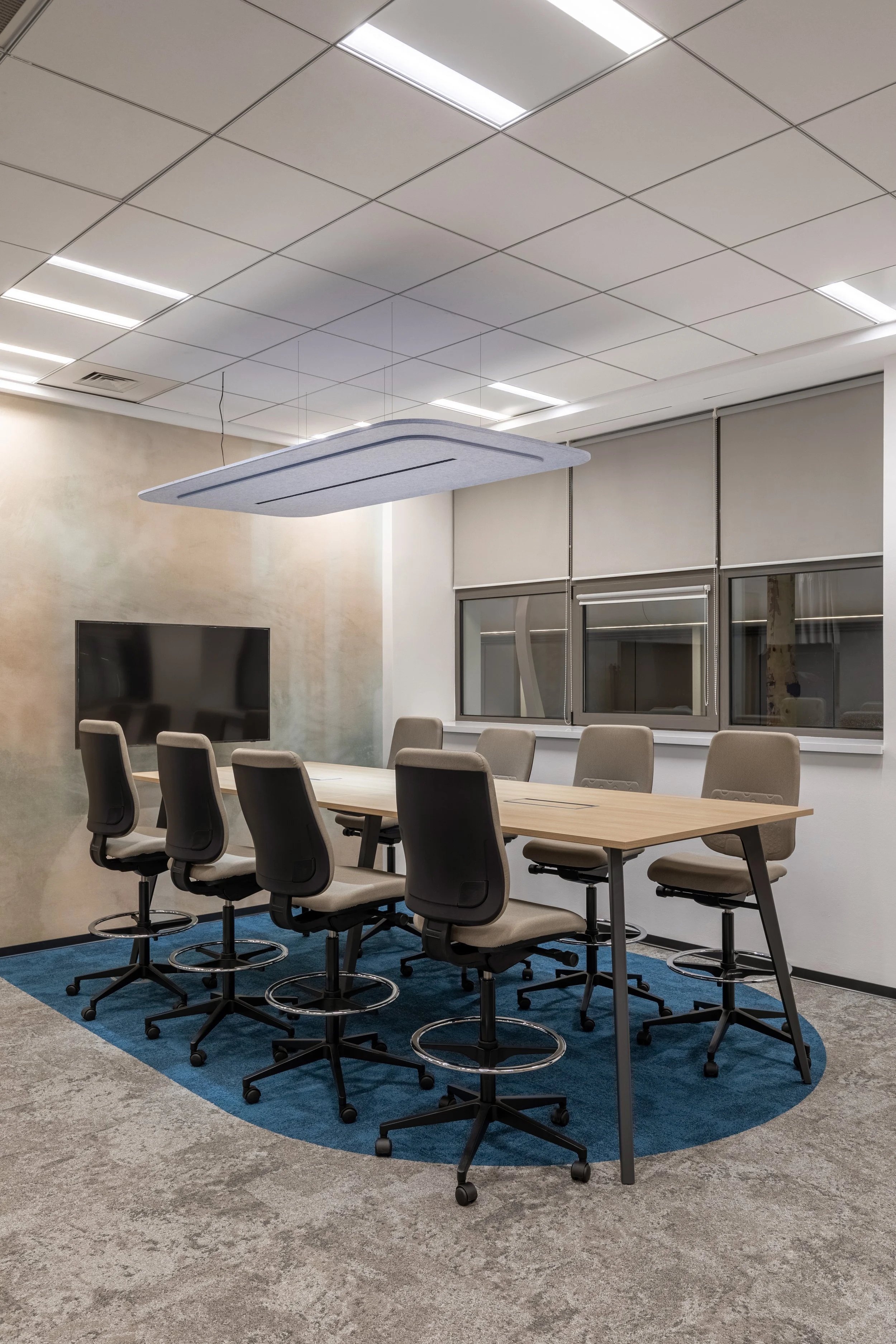 Empty modern conference room with a rectangular table, nine chairs, a wall-mounted flat screen, and windows with blinds.