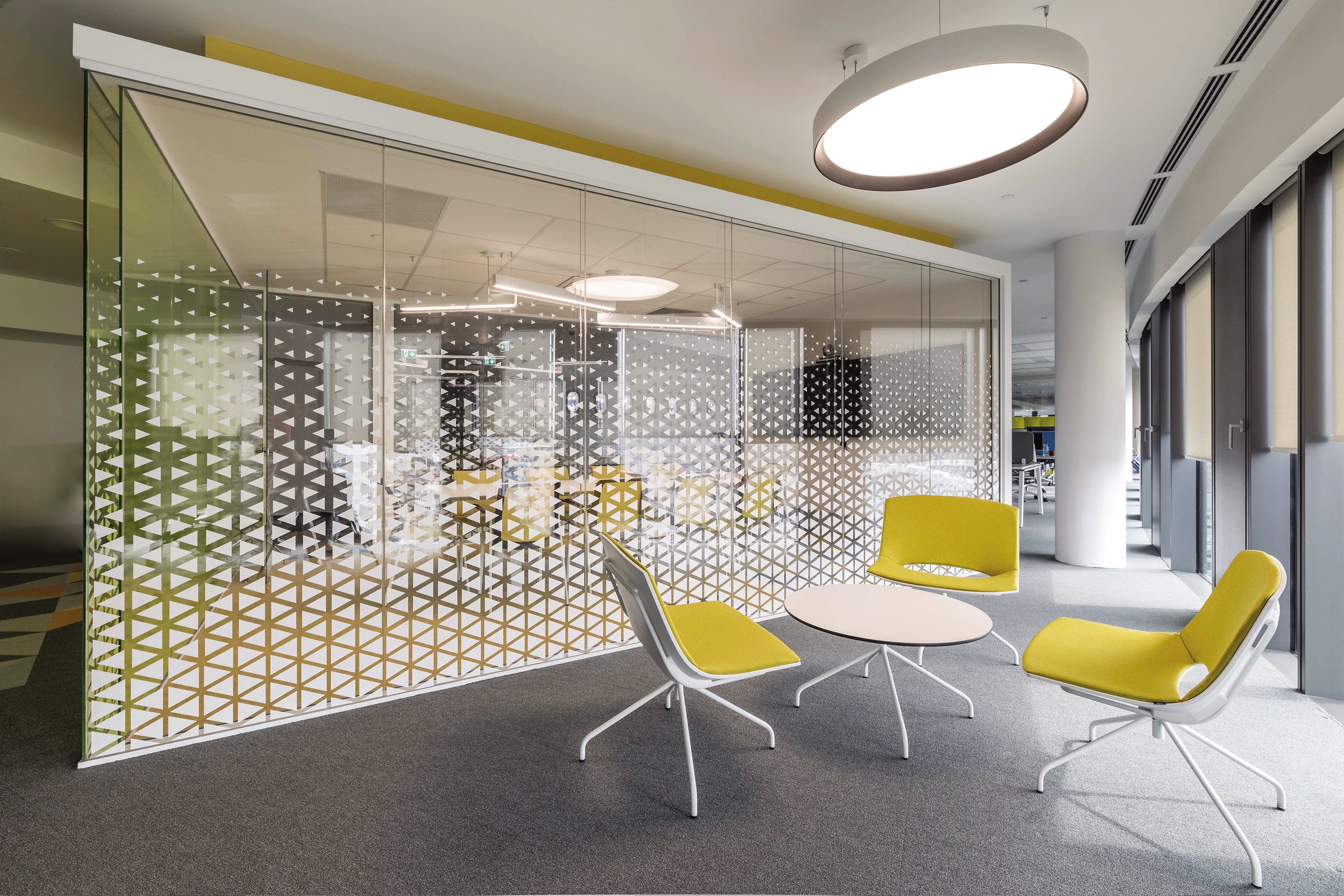 Modern office lounge area with yellow cushioned chairs around a small white round table, separated by a glass partition with a geometric pattern, and large windows along the right wall allowing natural light.