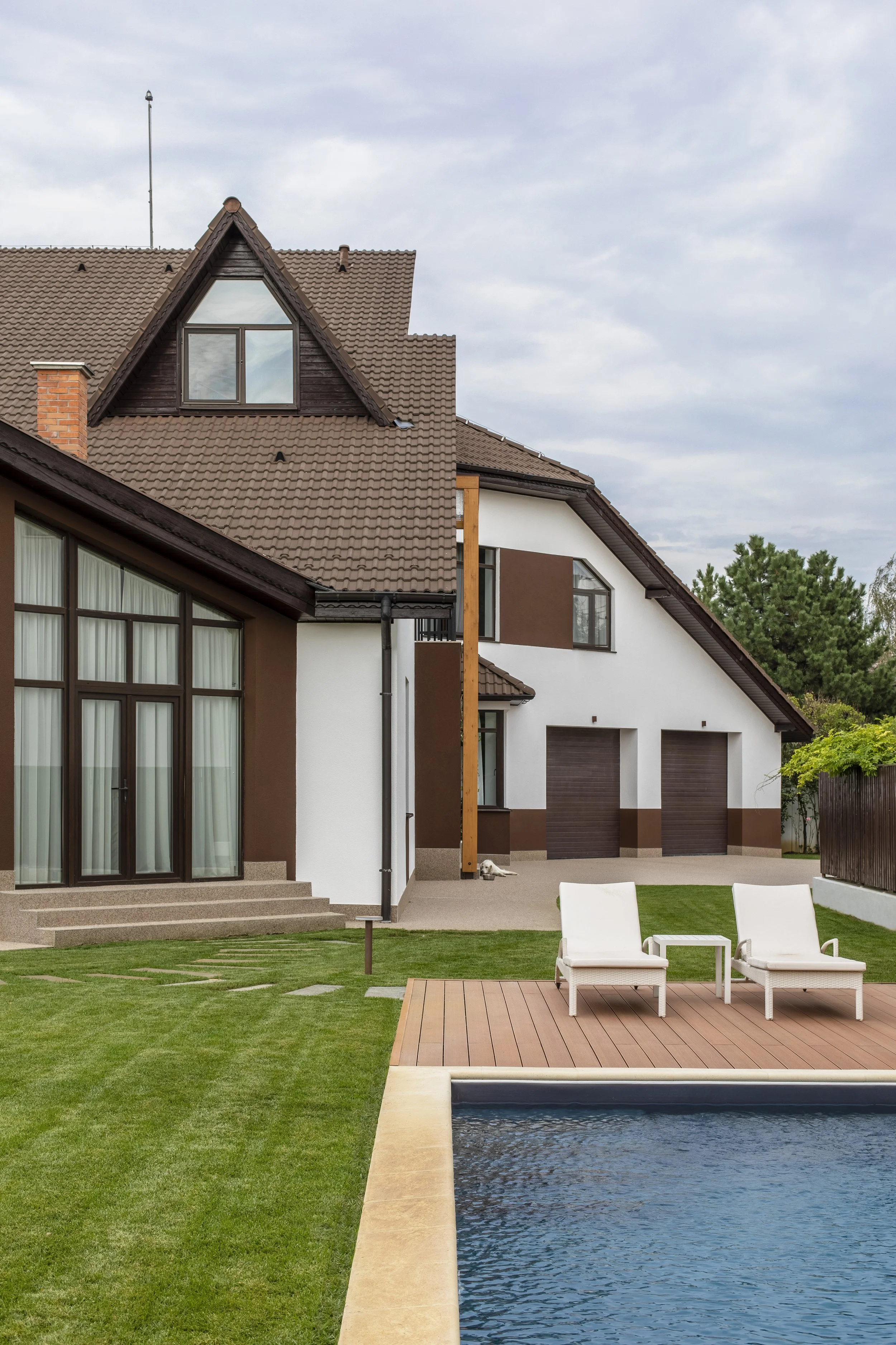 Modern house with a backyard pool, two lounge chairs, green lawn, and cloudy sky.