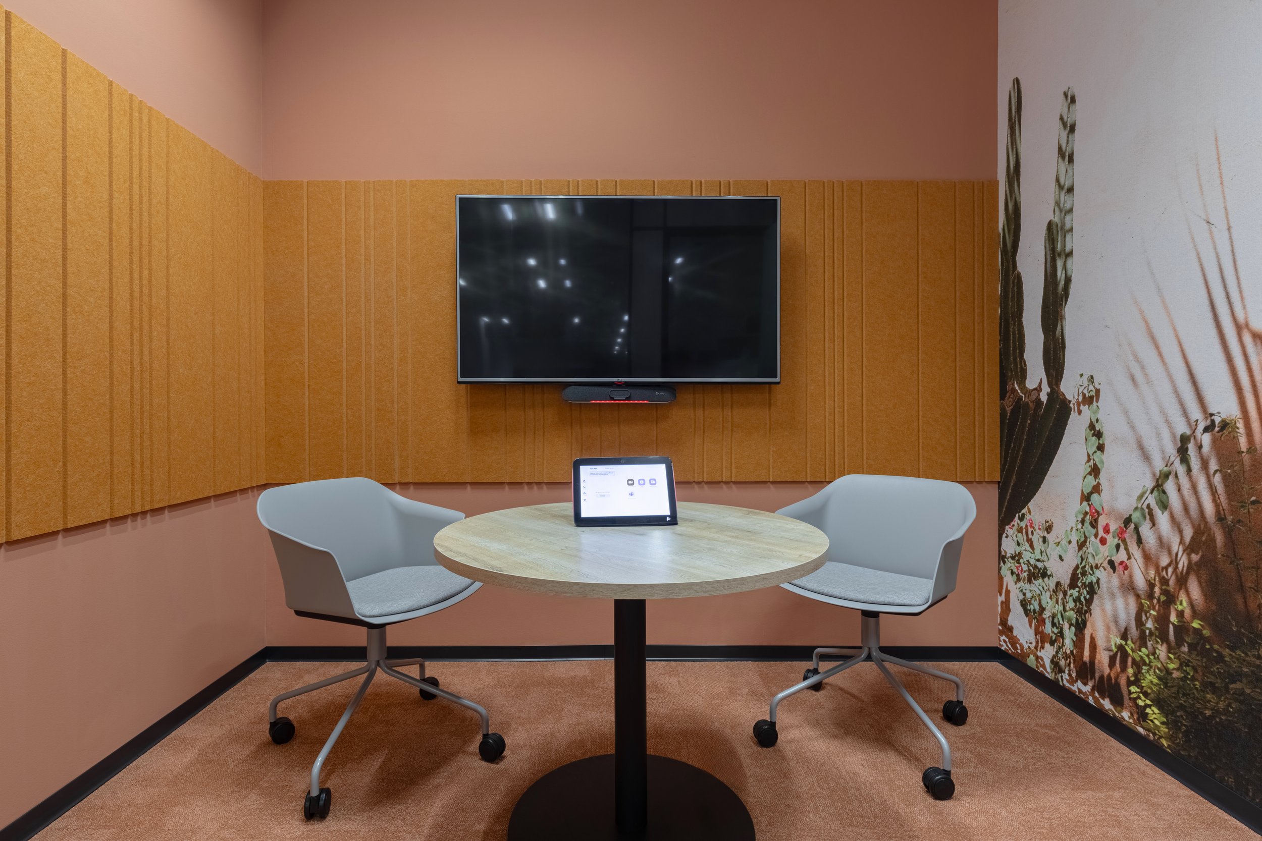 A small conference room with two white chairs around a round wooden table, a tablet on the table, an orange wall with soundproof panels, and a large flat-screen TV mounted on the wall.