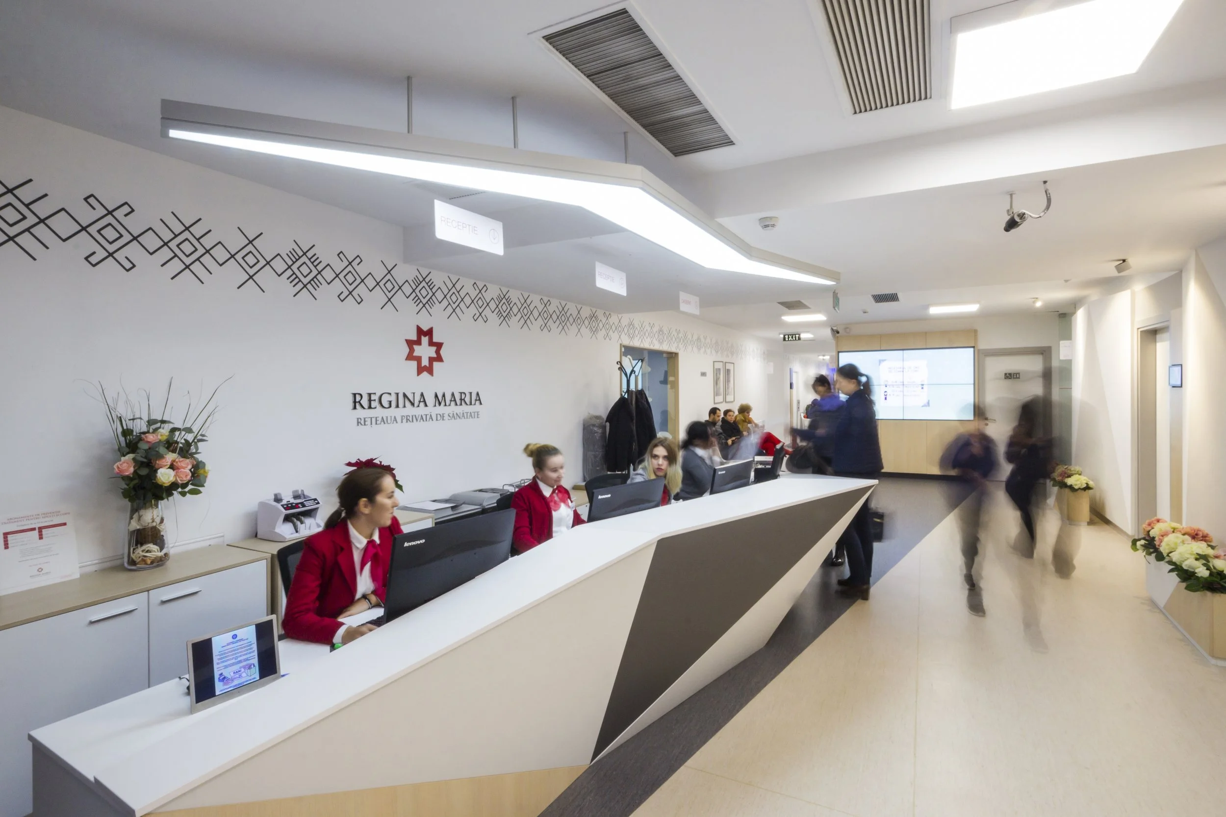 Reception area at Regina Maria healthcare facility with staff members working at the counter, patients and visitors walking by, and a sign displaying the Regina Maria logo and name on the wall, along with decorative floral arrangements and directional signs.