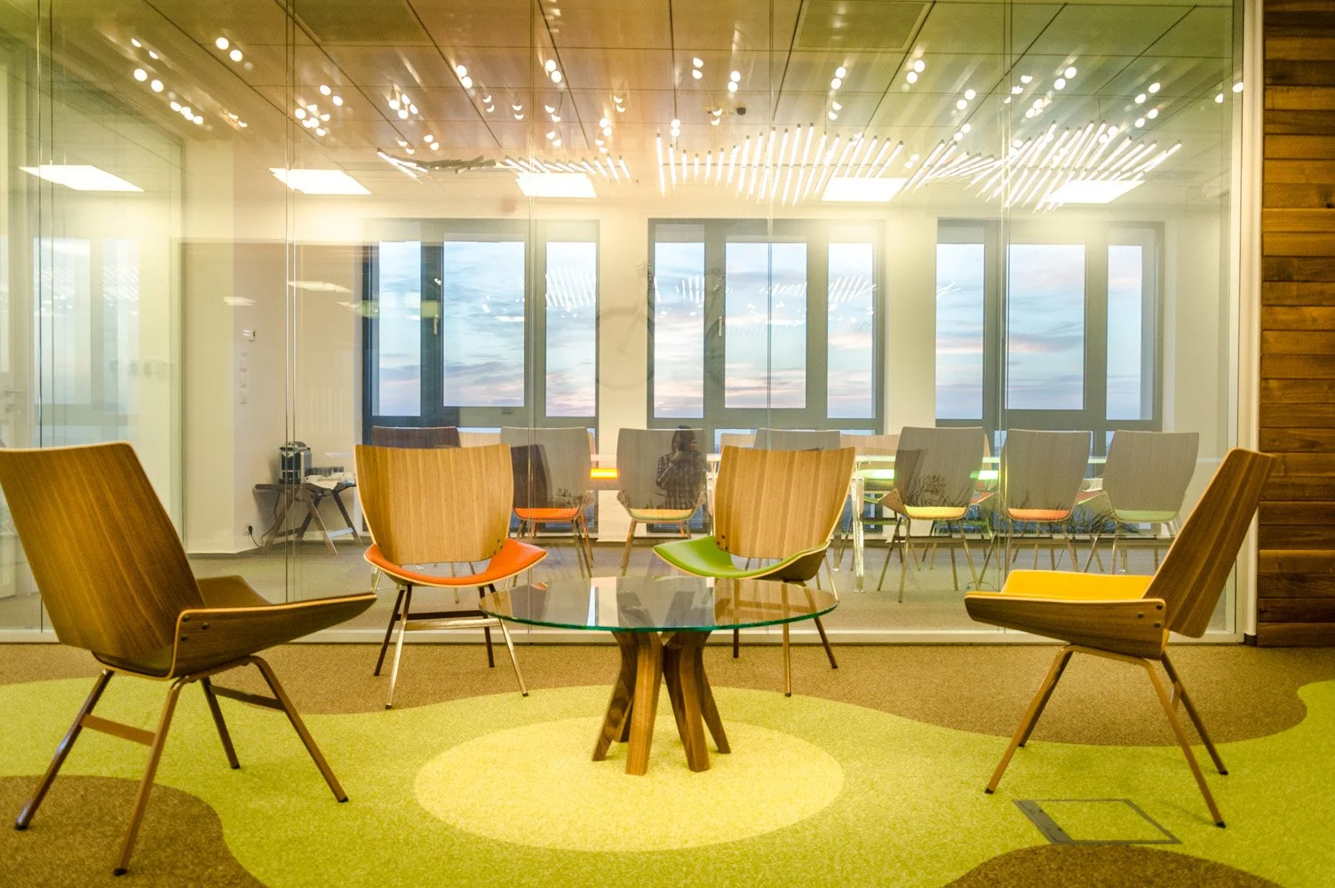 Modern conference room with glass walls, wooden chairs, and a glass table, overlooking a sunset sky outside.