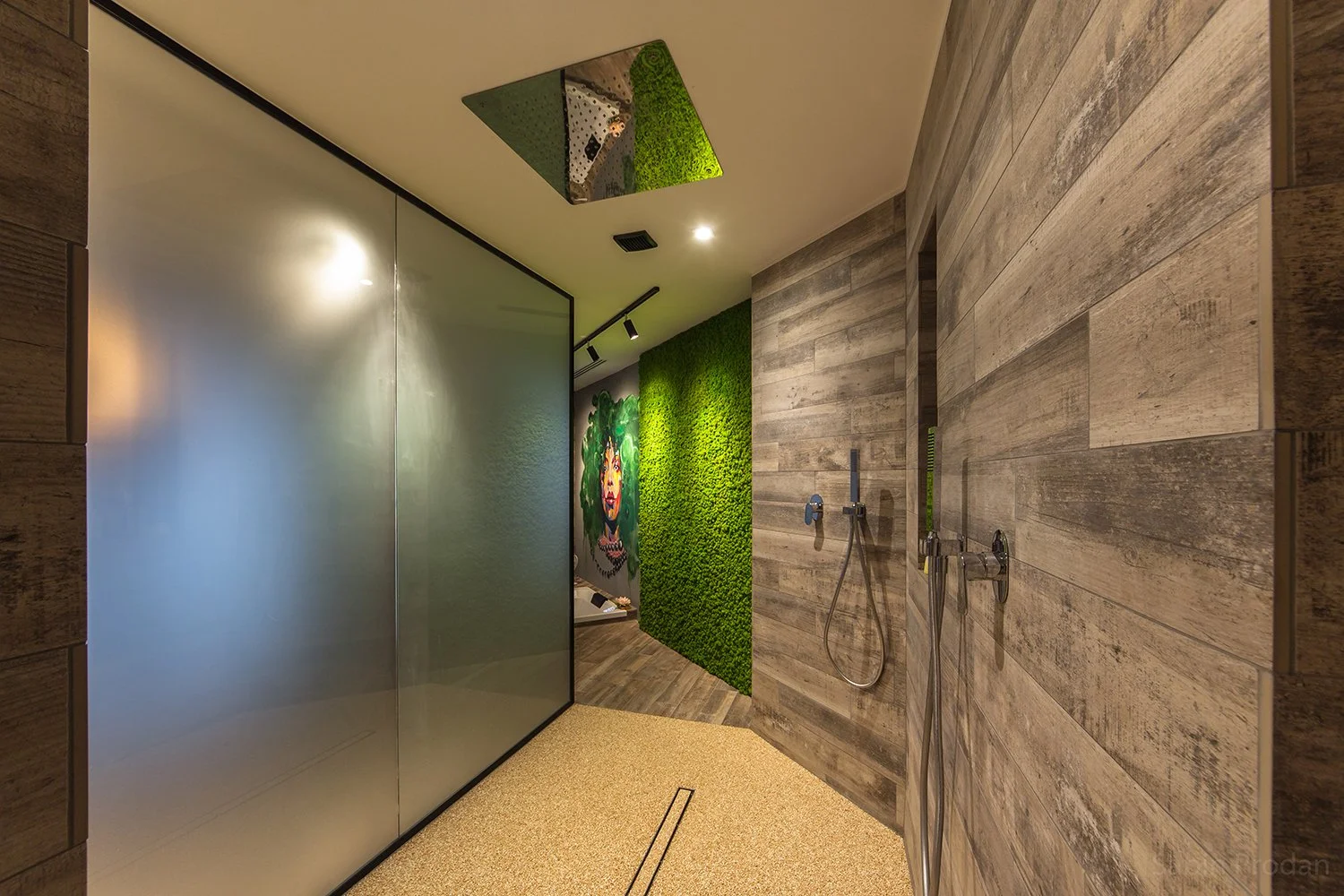 Modern shower area with wooden wall panels, a green moss wall with an artistic mural of a woman's face, a frosted glass partition, and rain shower fixtures.